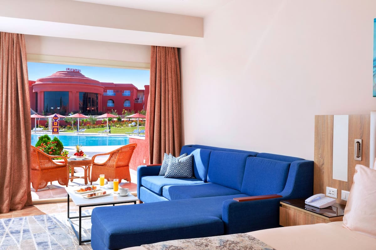 A hotel room with a blue sofa, a dining table with orange chairs, a view of a pool and a red building outside.