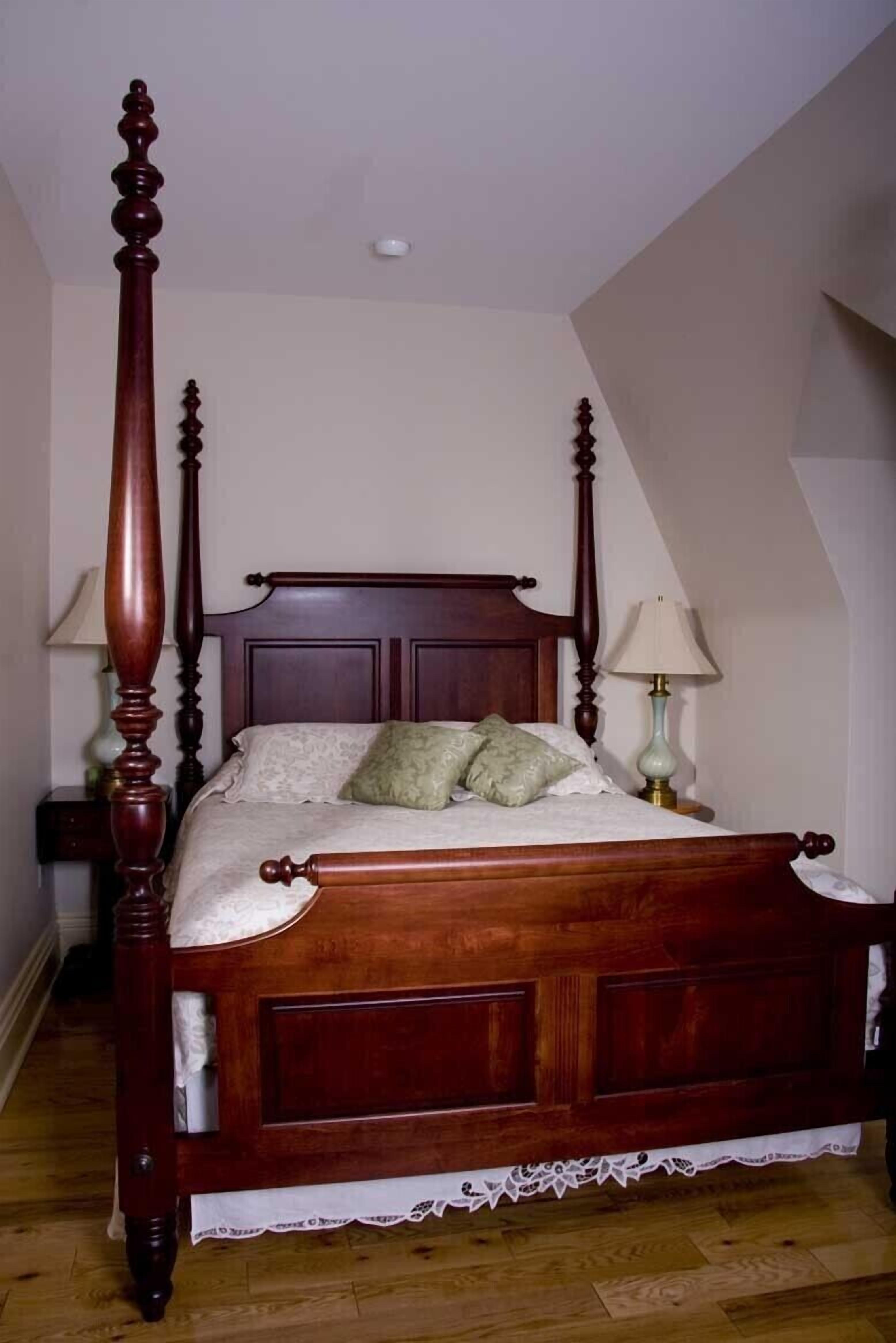 Uma cama de dossel com peseira e peitoril de madeira, saia de cama com rendas e duas luminárias de cabeceira.