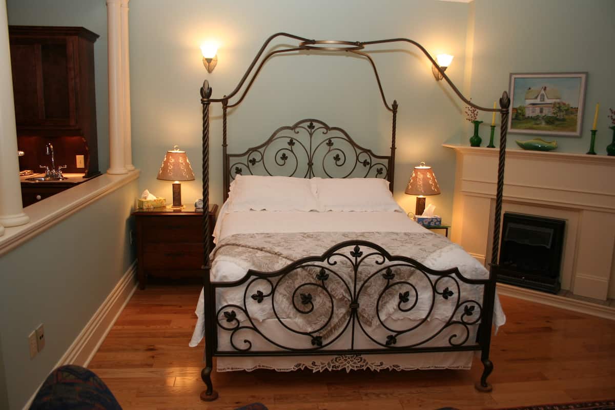 A four-poster bed with a lace bedspread, two bedside lamps, a nightstand with a lamp, a fireplace, and a painting on the wall.