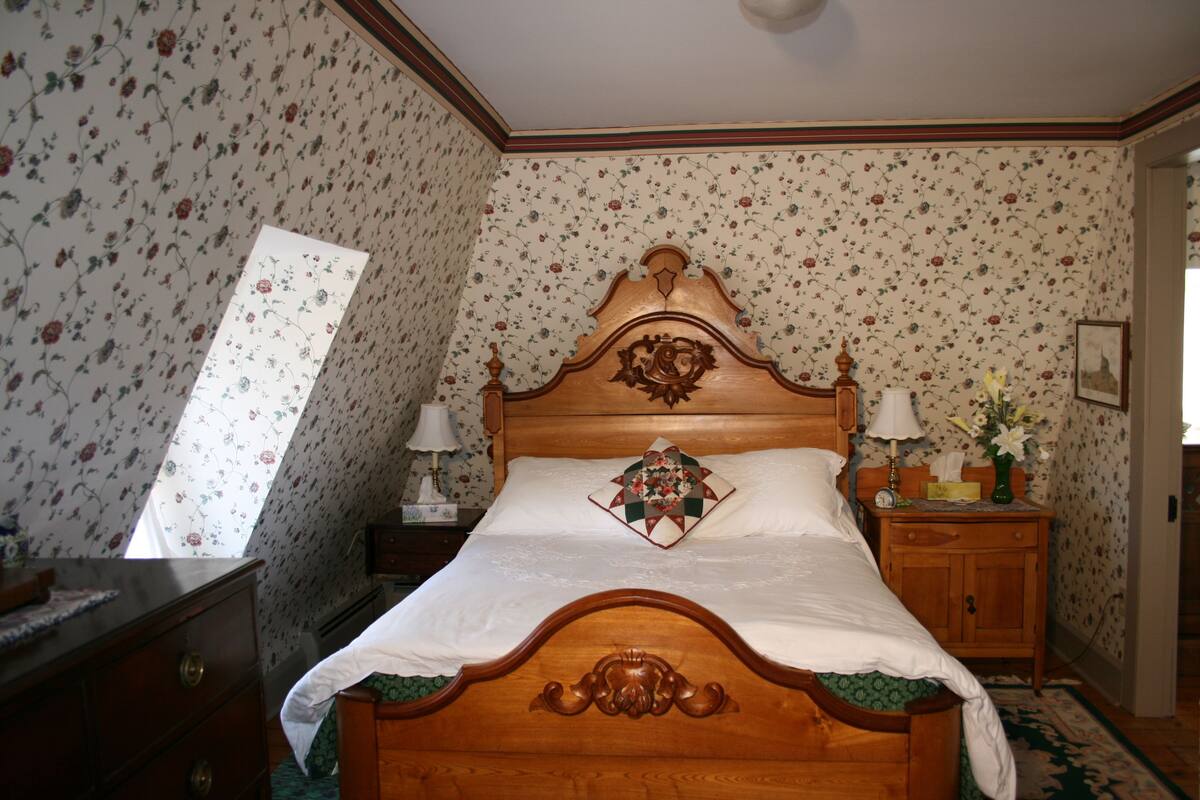 A bedroom with a wooden bed, floral wallpaper, a nightstand with a lamp, and a dresser.