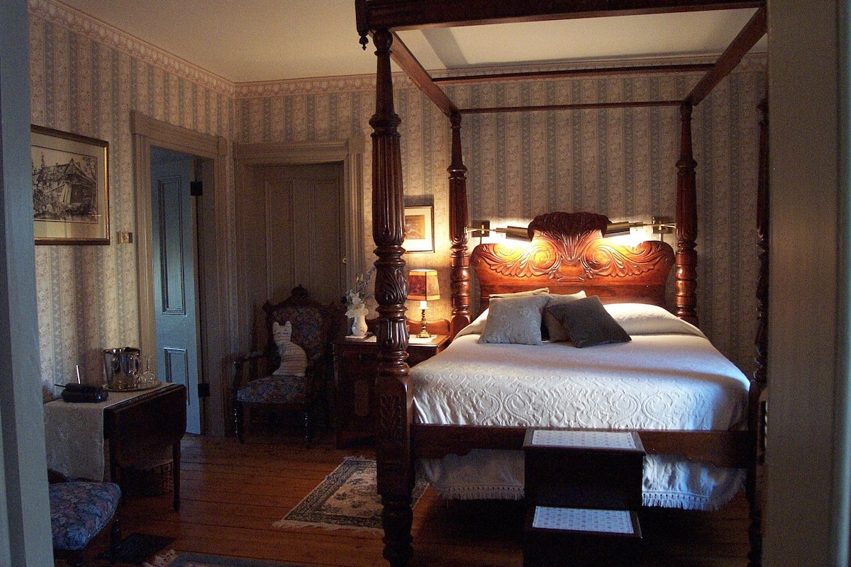 A four-poster bed with a wooden headboard, a bedside table with a lamp, a chair, and a small table with a vase.