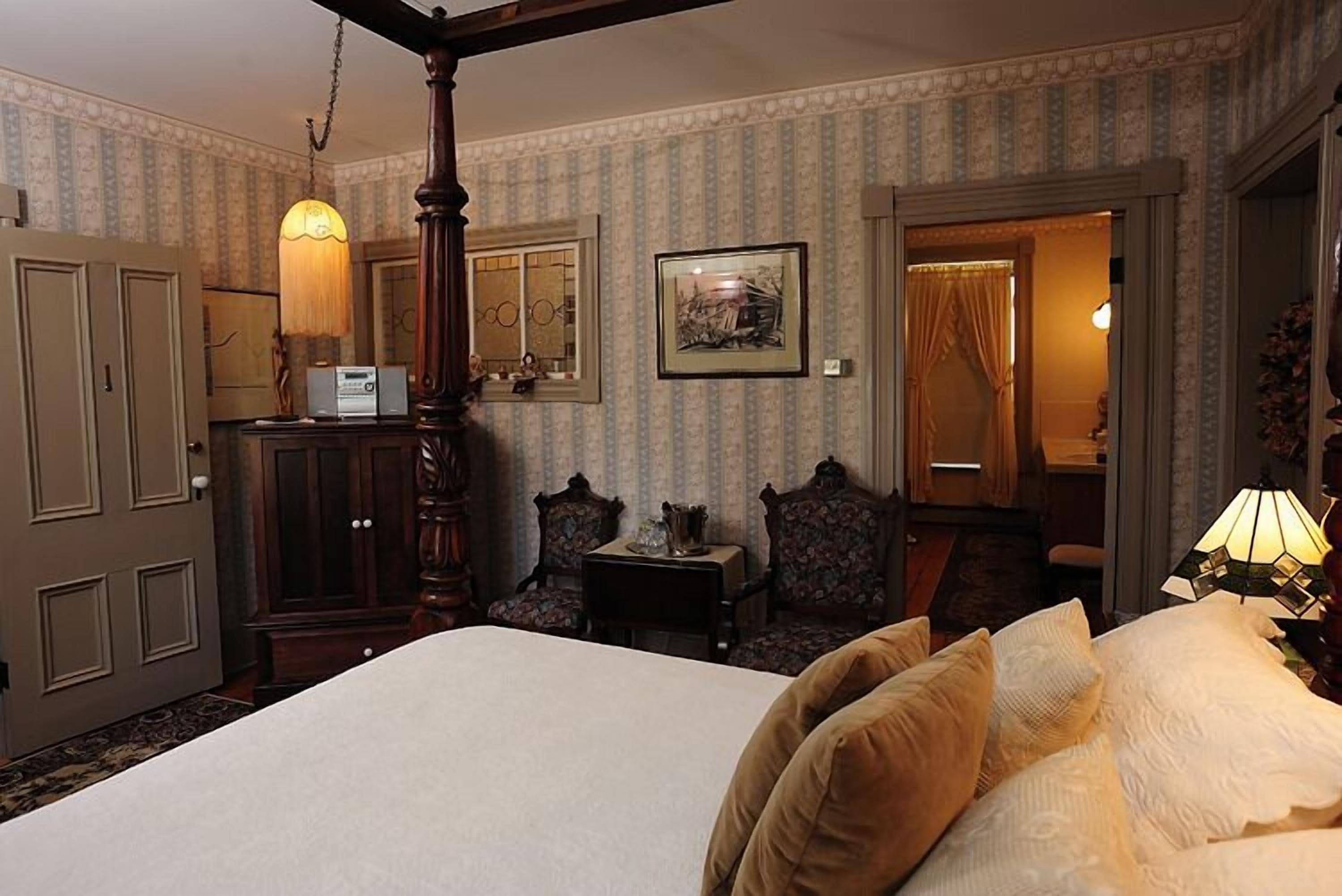 A traditional hotel room with a four-poster bed, a wooden wardrobe, a small table with a lamp, and a framed picture on the wall.
