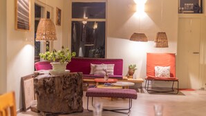 Una sala de estar acogedora con un sofá, un reposapiés y una mesa de centro. Hay luces colgantes y una ventana con cortinas.