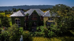 Una casa con techo pronunciado, rodeada de abundante vegetación y un paisaje montañoso al fondo.