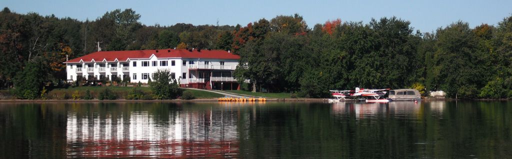 Un grand bâtiment blanc, surmonté d’un toit rouge, situé au bord d’un lac, entouré d’arbres et doté d’un quai où sont amarrés des bateaux.