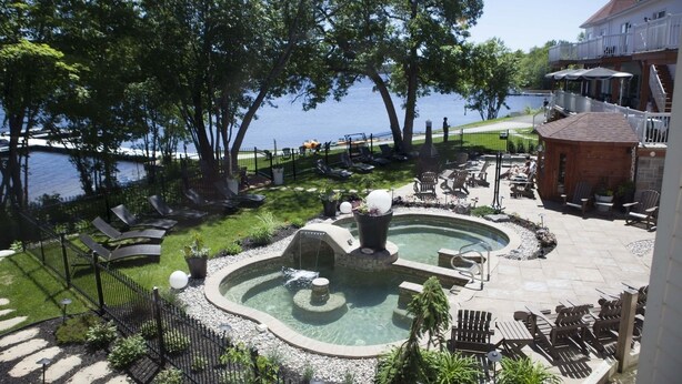 A pool area with lounge chairs and a hot tub, surrounded by trees and a view of the water. - Manoir du Lac William (St-Ferdinand)