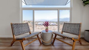 A room with two woven chairs, a round table with a potted plant, and a large window with a view of a desert landscape.
