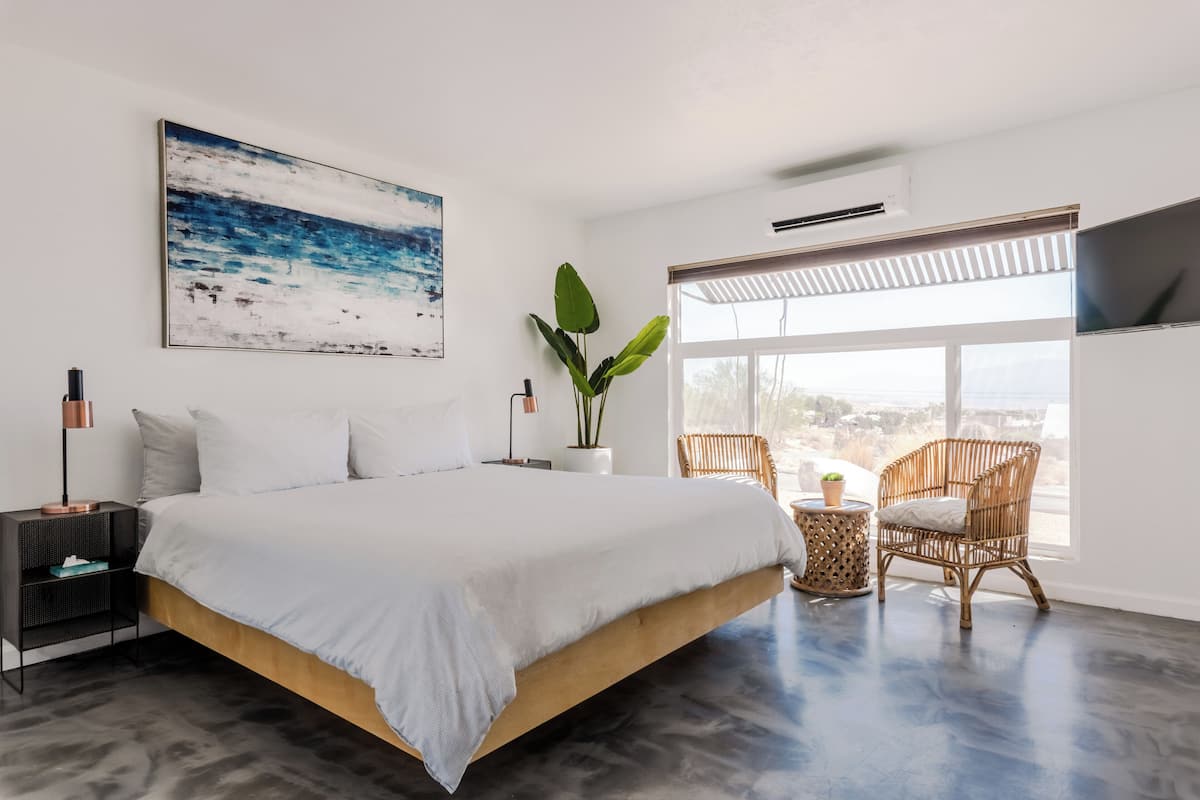A modern bedroom with a large bed, a view of the city, a potted plant, and a wicker chair.