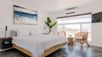 A modern bedroom with a large bed, a view of the city, a potted plant, and a wicker chair.