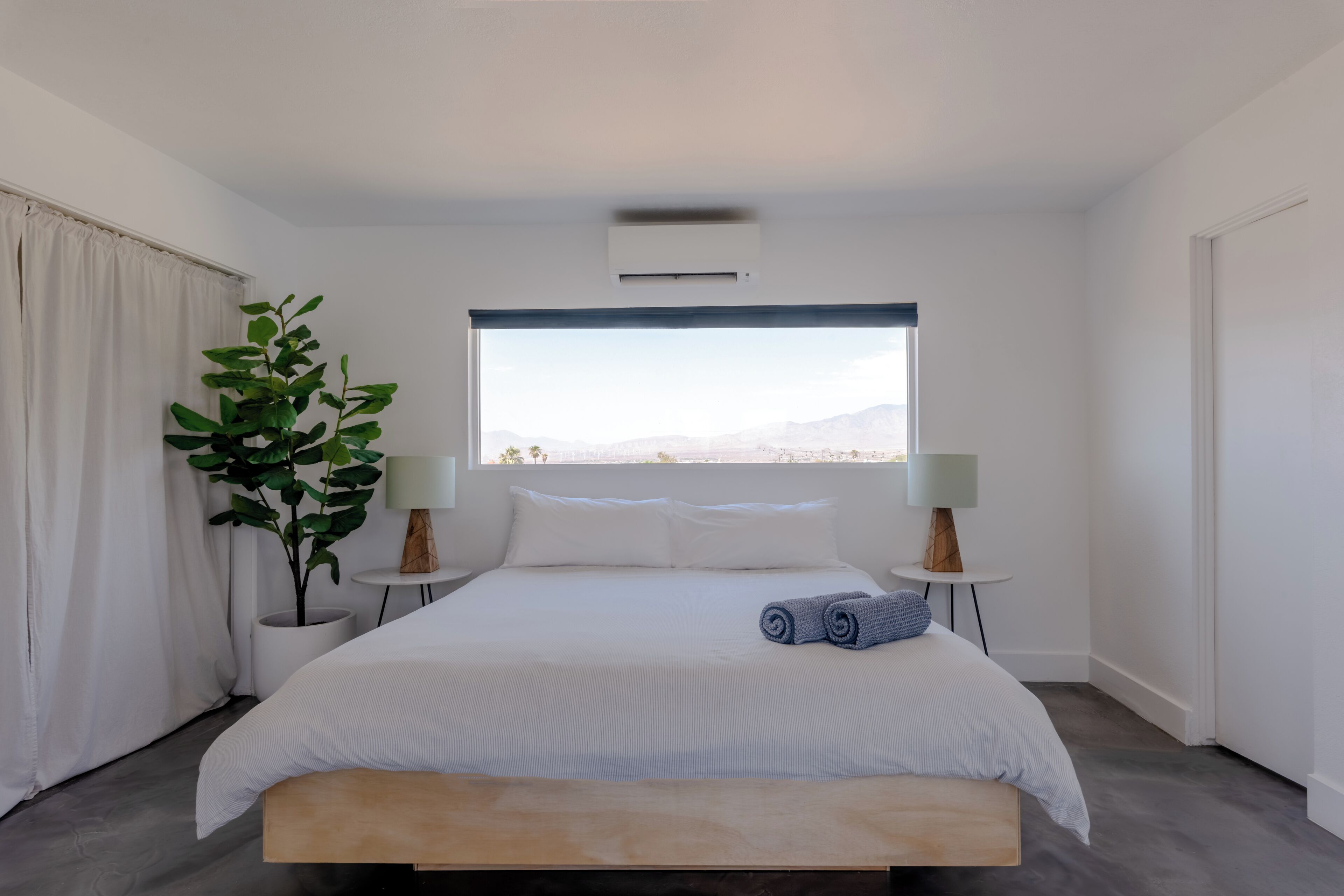 Une chambre moderne avec un grand lit, une plante en pot, deux lampes de chevet et une vue sur les montagnes par la fenêtre.