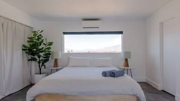 Une chambre moderne avec un grand lit, une plante en pot, deux lampes de chevet et une vue sur les montagnes par la fenêtre.