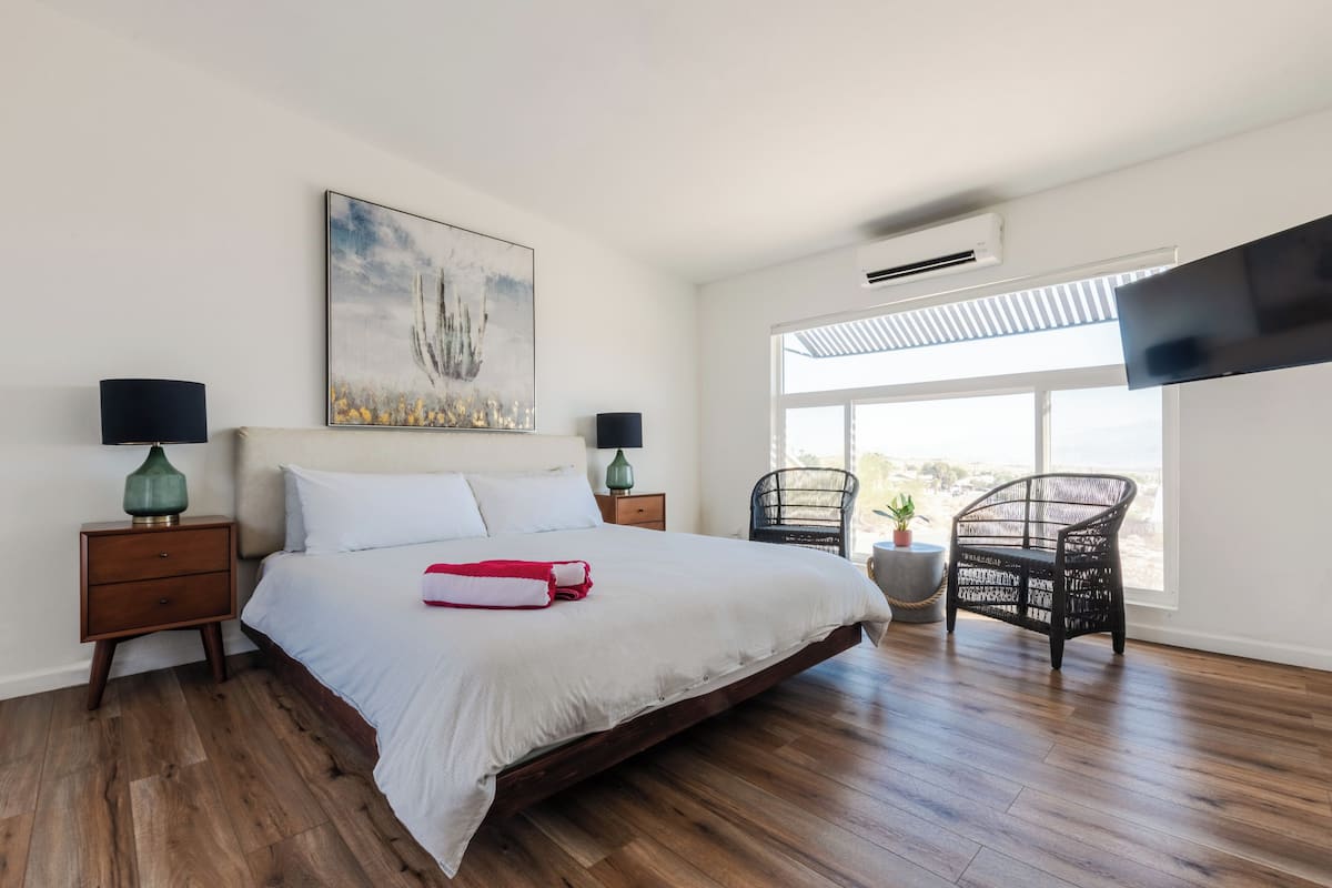 A bedroom with a large bed, two nightstands, a wall-mounted TV, a window with a view, and a ceiling fan.