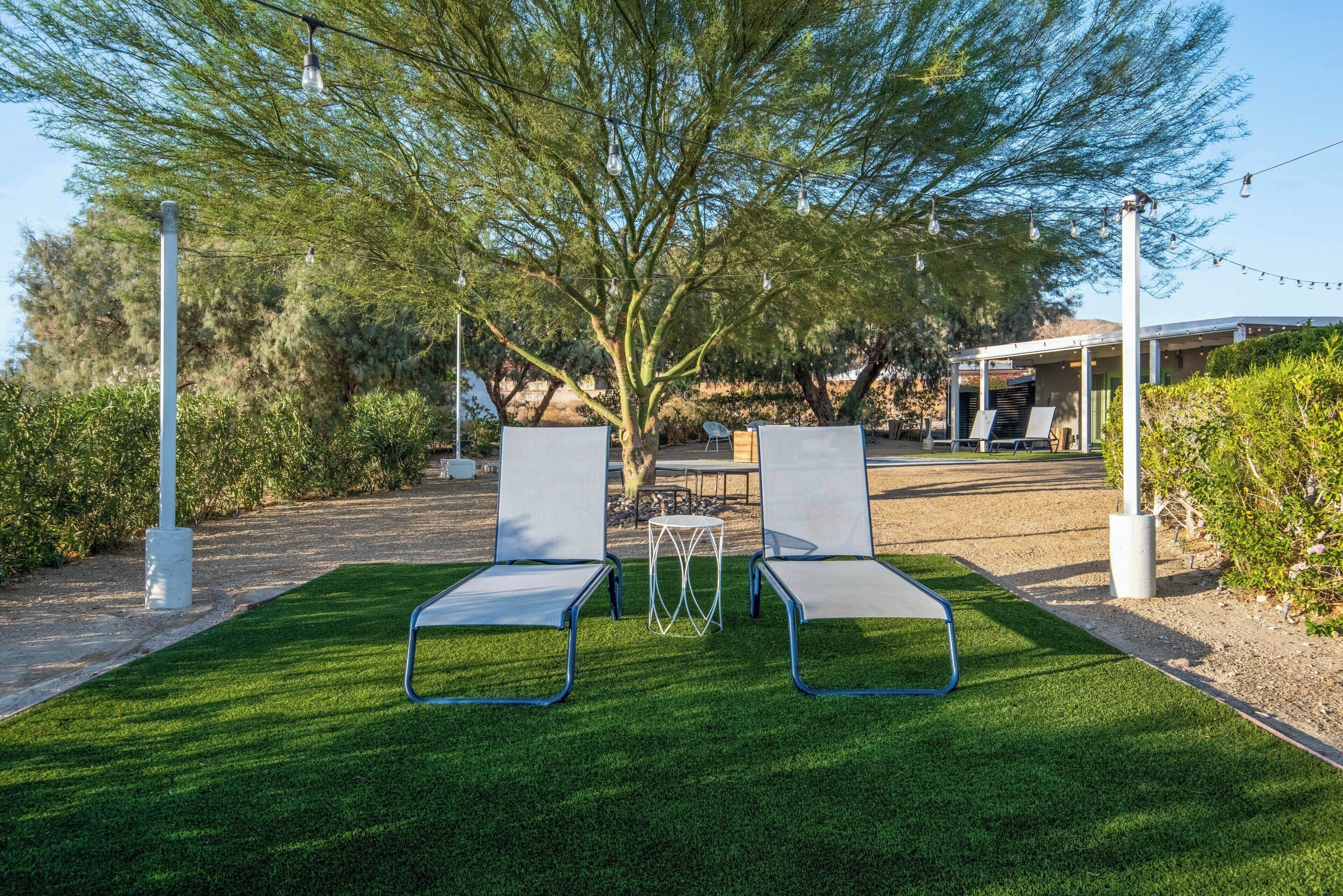 Two lounge chairs on a well-manicured lawn with a pergola overhead, surrounded by greenery and a building in the background.