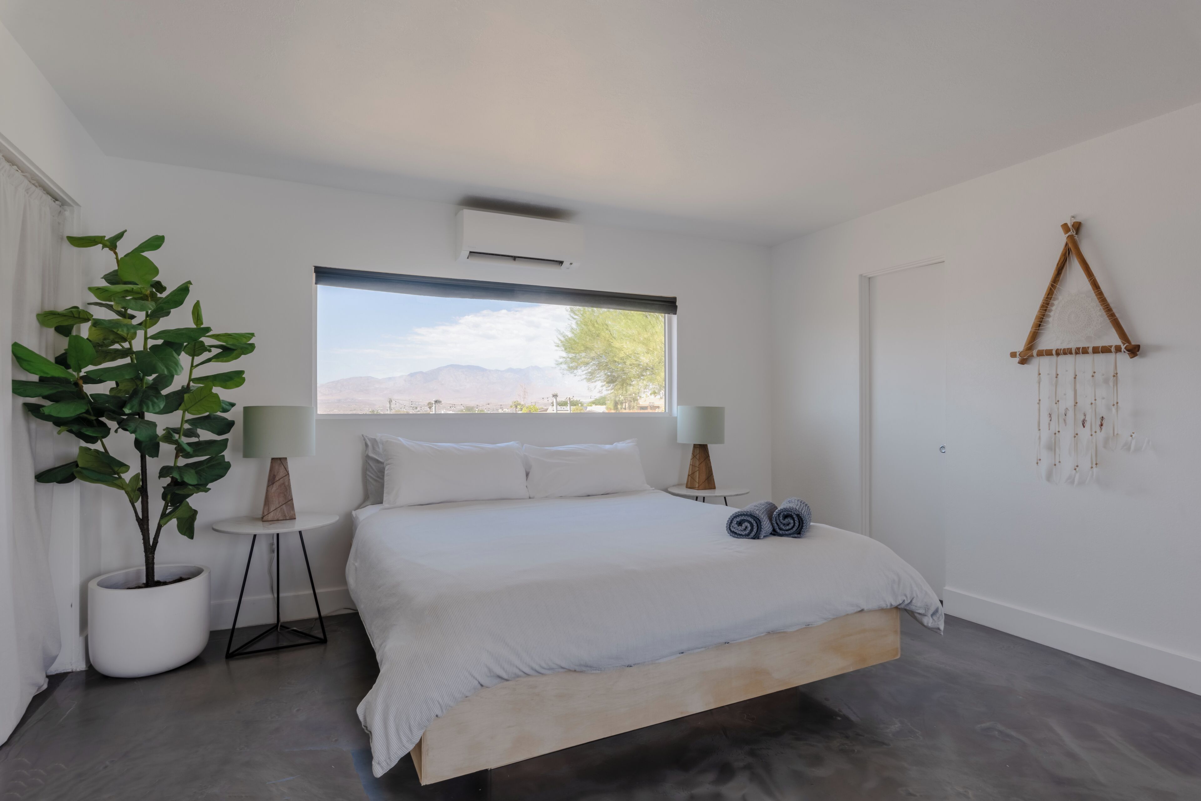 A modern bedroom with a large bed, a potted plant, two bedside lamps, and a view of the outdoors.