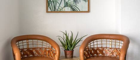 Deux fauteuils en osier avec des coussins en cuir, une petite table d’appoint en bois sur laquelle est posée une plante en pot, et un tableau encadré représentant une feuille tropicale accroché au mur.