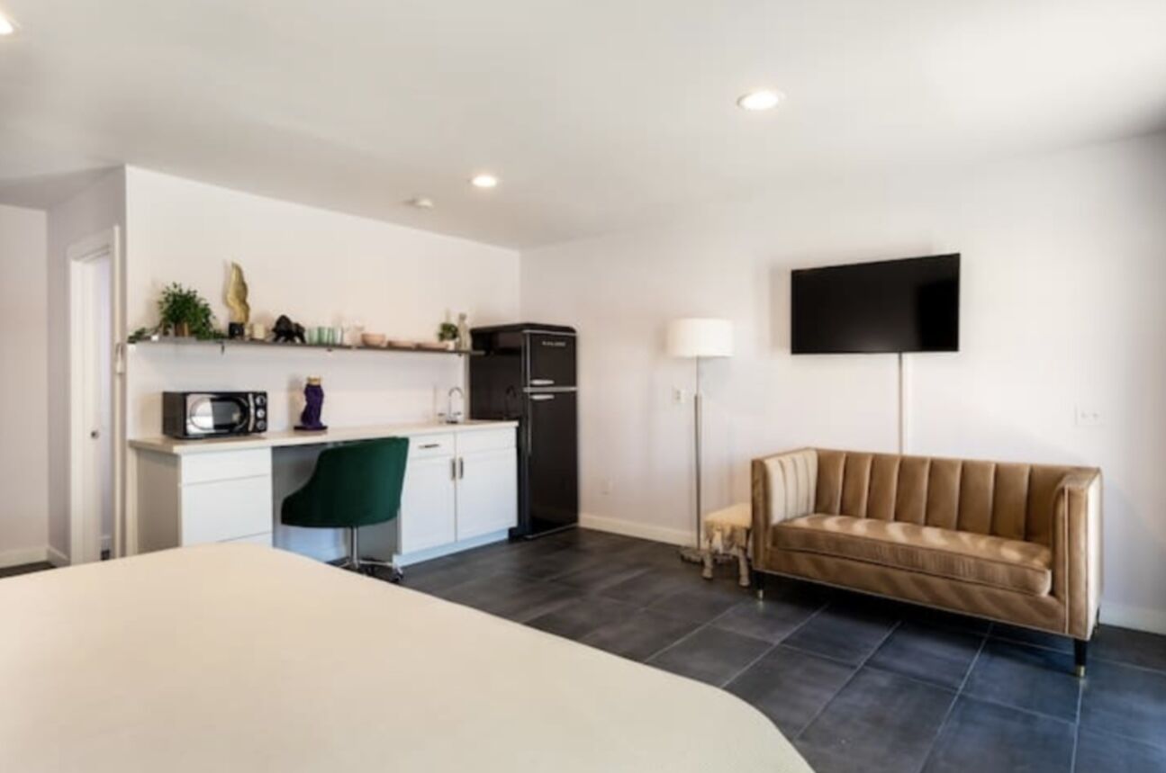 En moderne, minimalistisk stue med sofa, fladskærms-tv, tekøkken med mikrobølgeovn, køleskab og et lille spisebord.