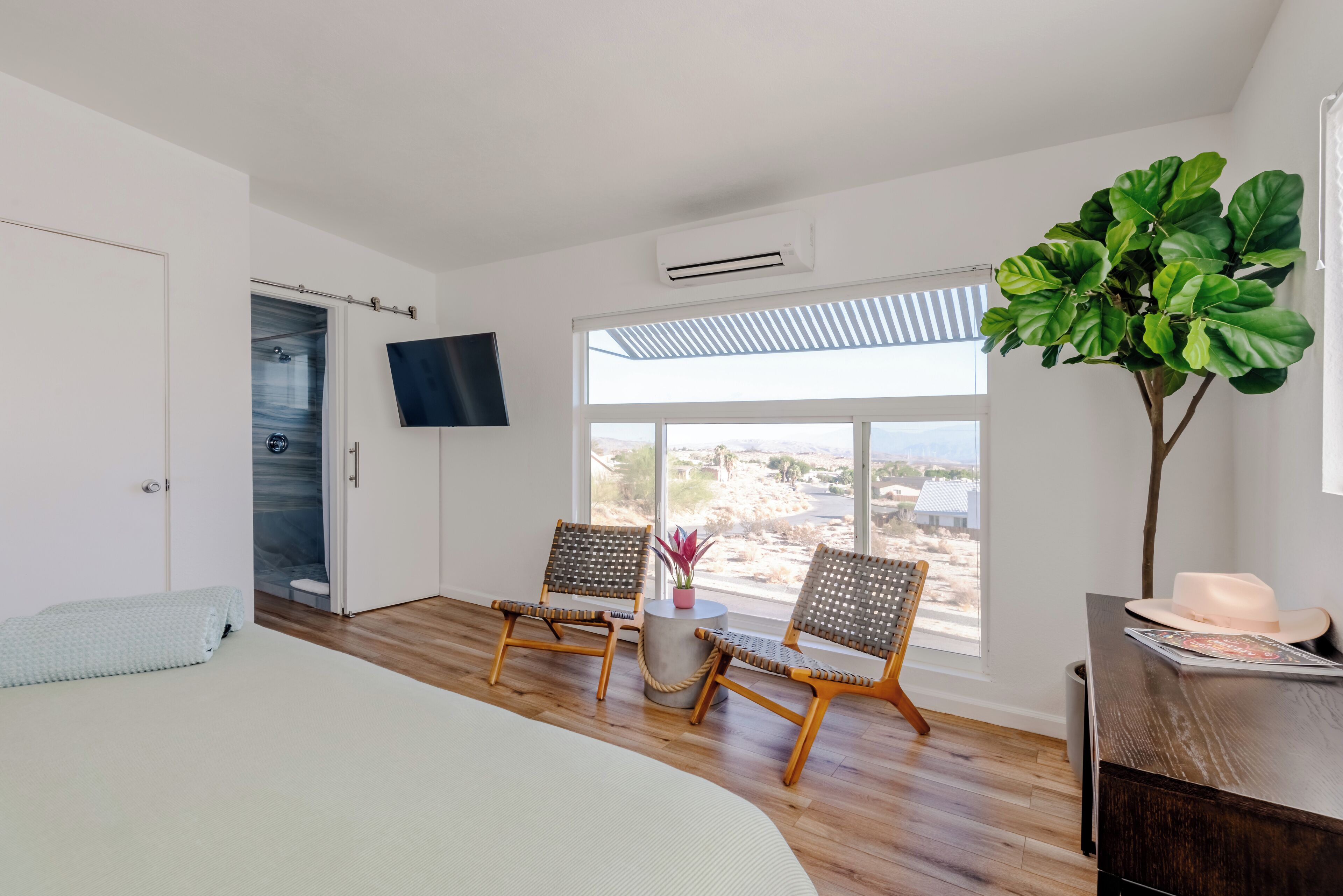 A bedroom with a bed, two wicker chairs, a small round table with a plant, a large potted plant, a TV mounted on the wall, and a view of the outdoors through a large window.