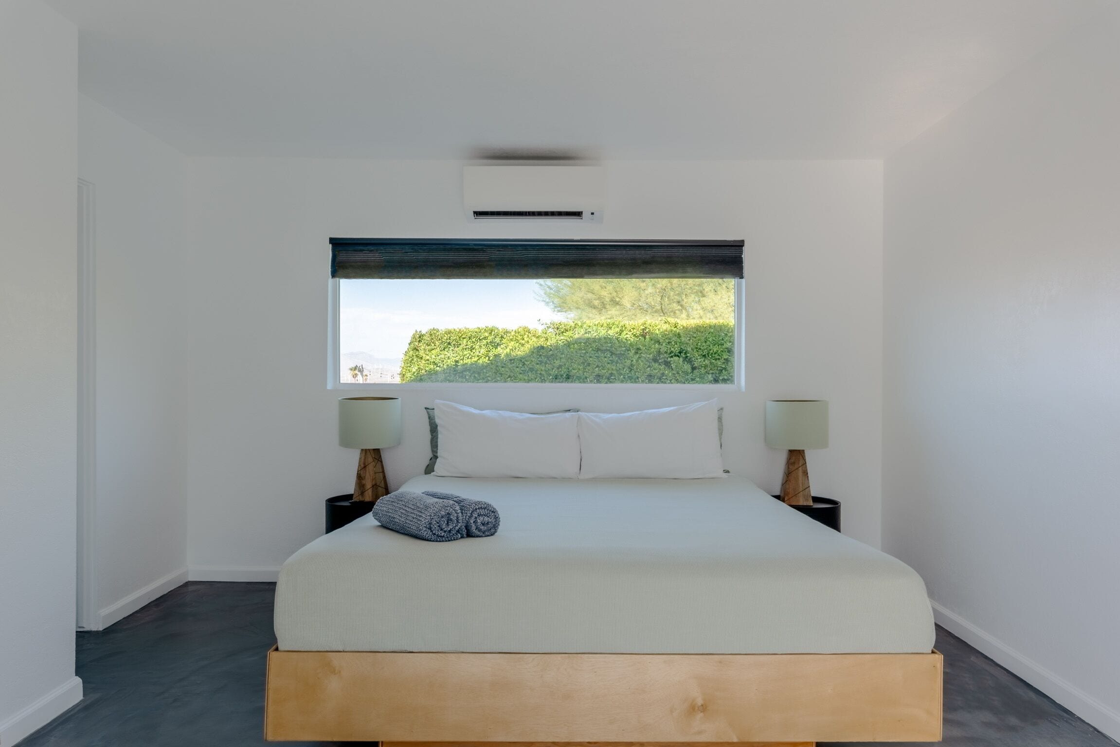 Un dormitorio con un ventanal grande, una cama con ropa de cama blanca, dos mesitas de noche con lámparas y un cabecero de madera.