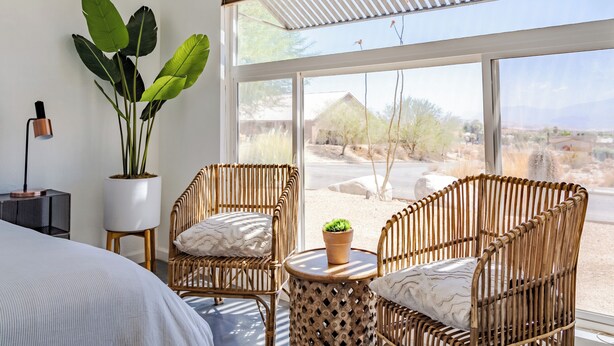 A room with wicker chairs, a potted plant, and a view of a desert landscape through large windows.