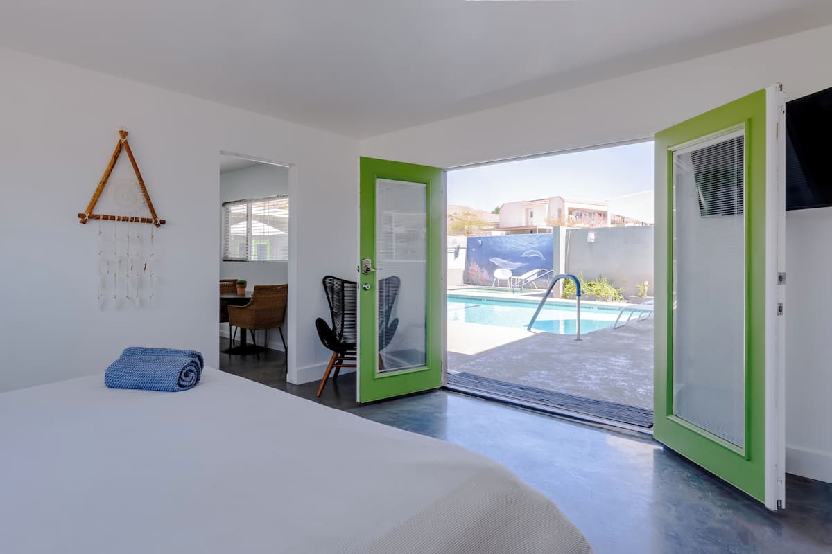 A modern hotel room with a bed, a green door leading to a pool area, and a view of the outside.