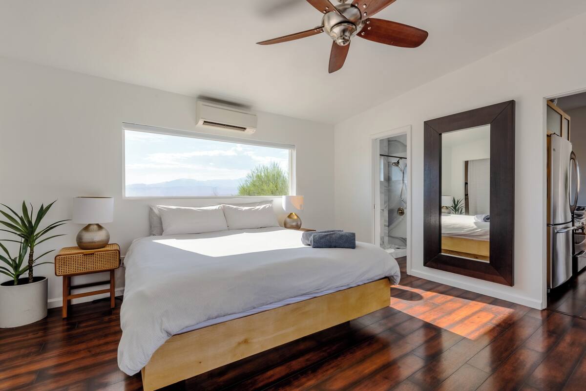 A modern bedroom with a large bed, wooden flooring, a ceiling fan, a large window with a mountain view, a bedside table with a lamp, and a mirror.