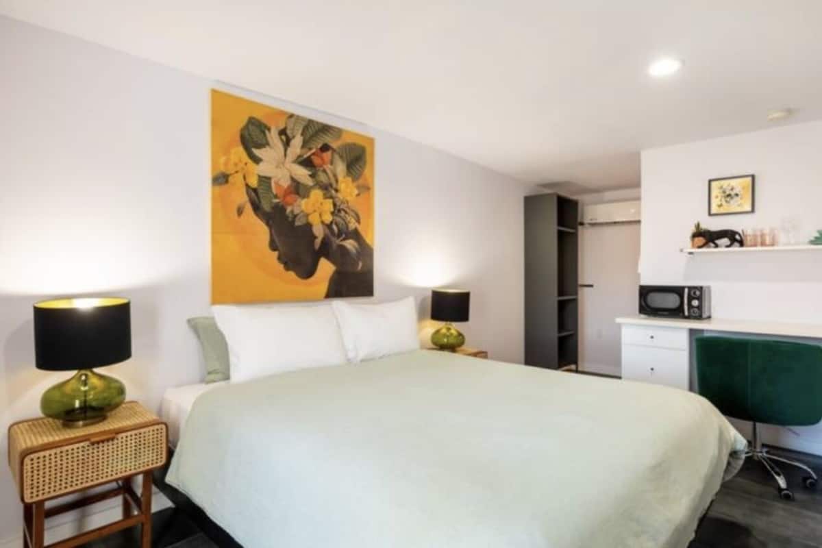 A hotel room with a bed, two bedside lamps, a green chair, a desk with a computer, a microwave, and a shelf with items.