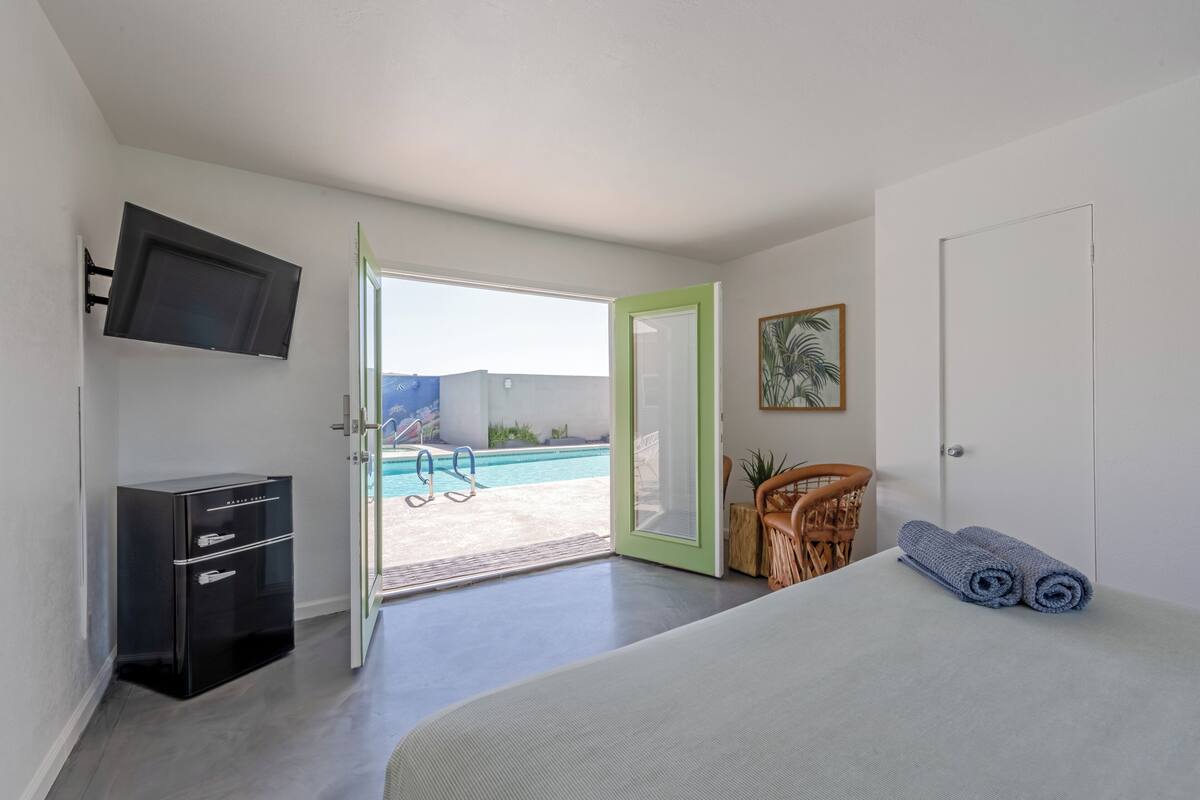 A bedroom with a bed, a television mounted on the wall, a wardrobe, a door leading to a pool area, and a chair.