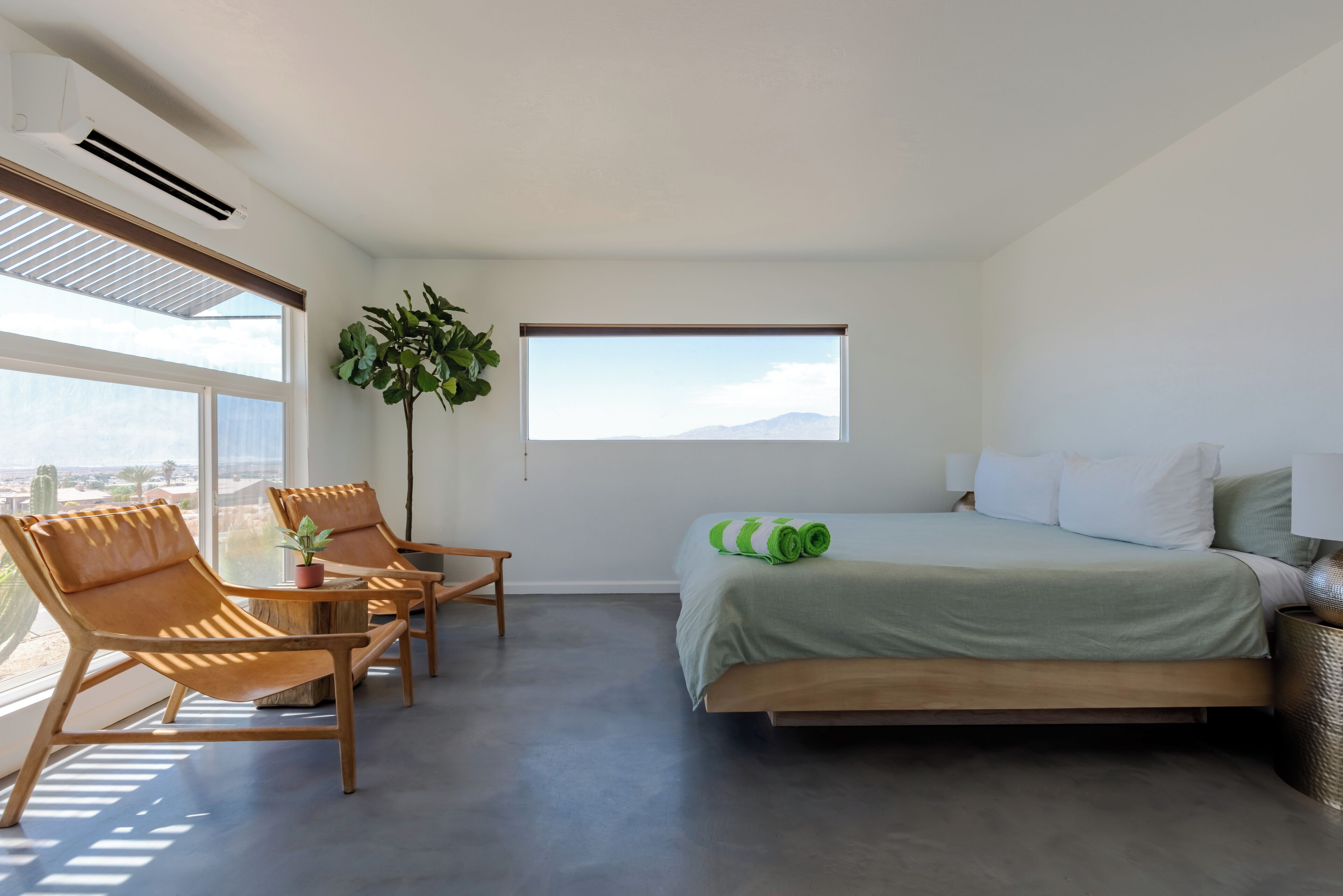 A modern bedroom with a bed, two leather chairs, a small table, a potted plant, and a large window with a view of the mountains.