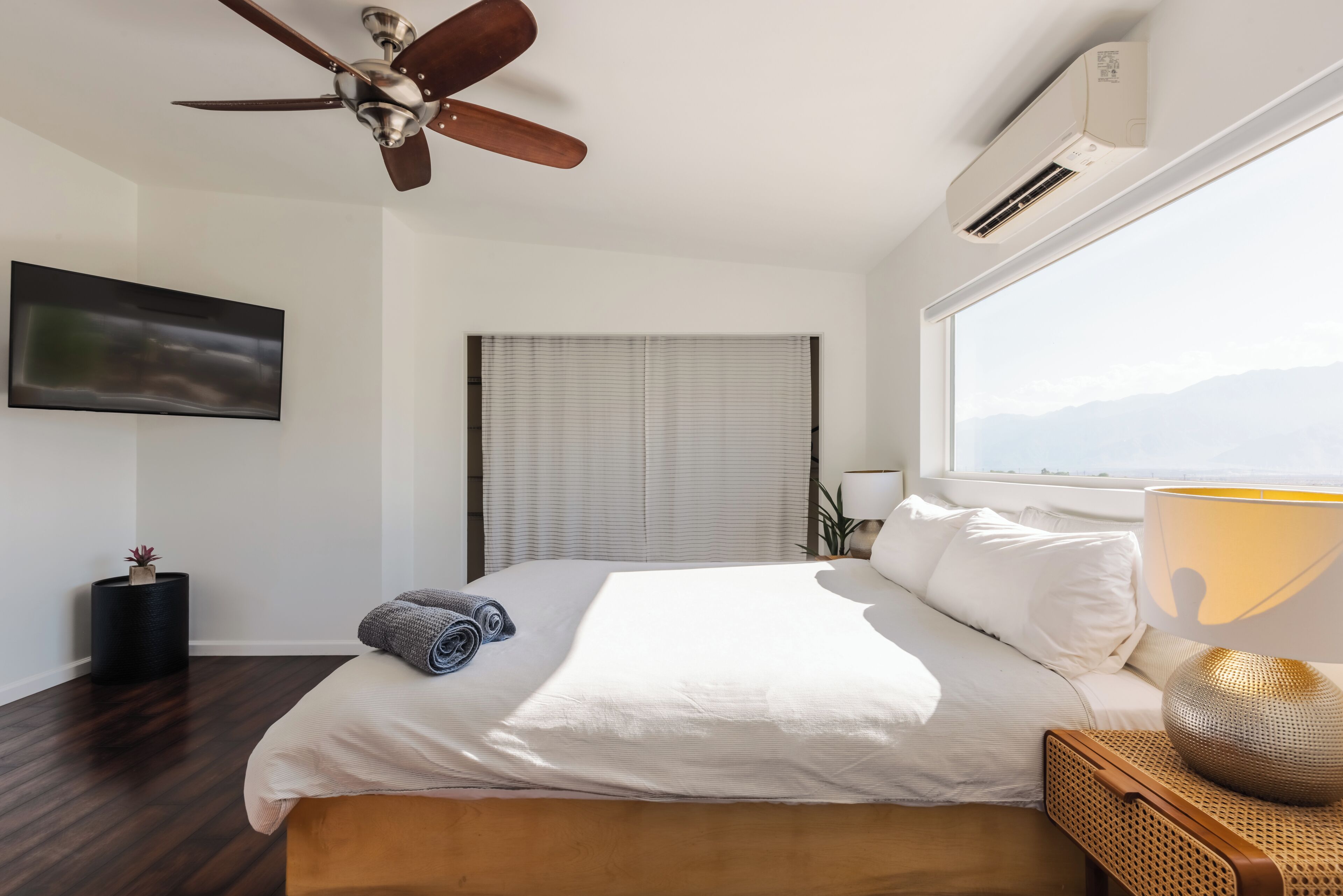 A modern bedroom with a large bed, ceiling fan, air conditioning unit, TV, and a window with a view.