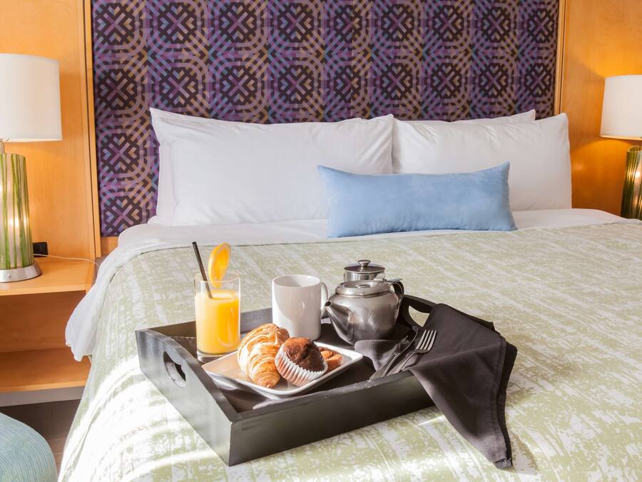 Ein Hotelzimmer mit einem ordentlich bezogenem Bett, einem Tablett mit Frühstücksangeboten und einem gemusterten Kopfteil.