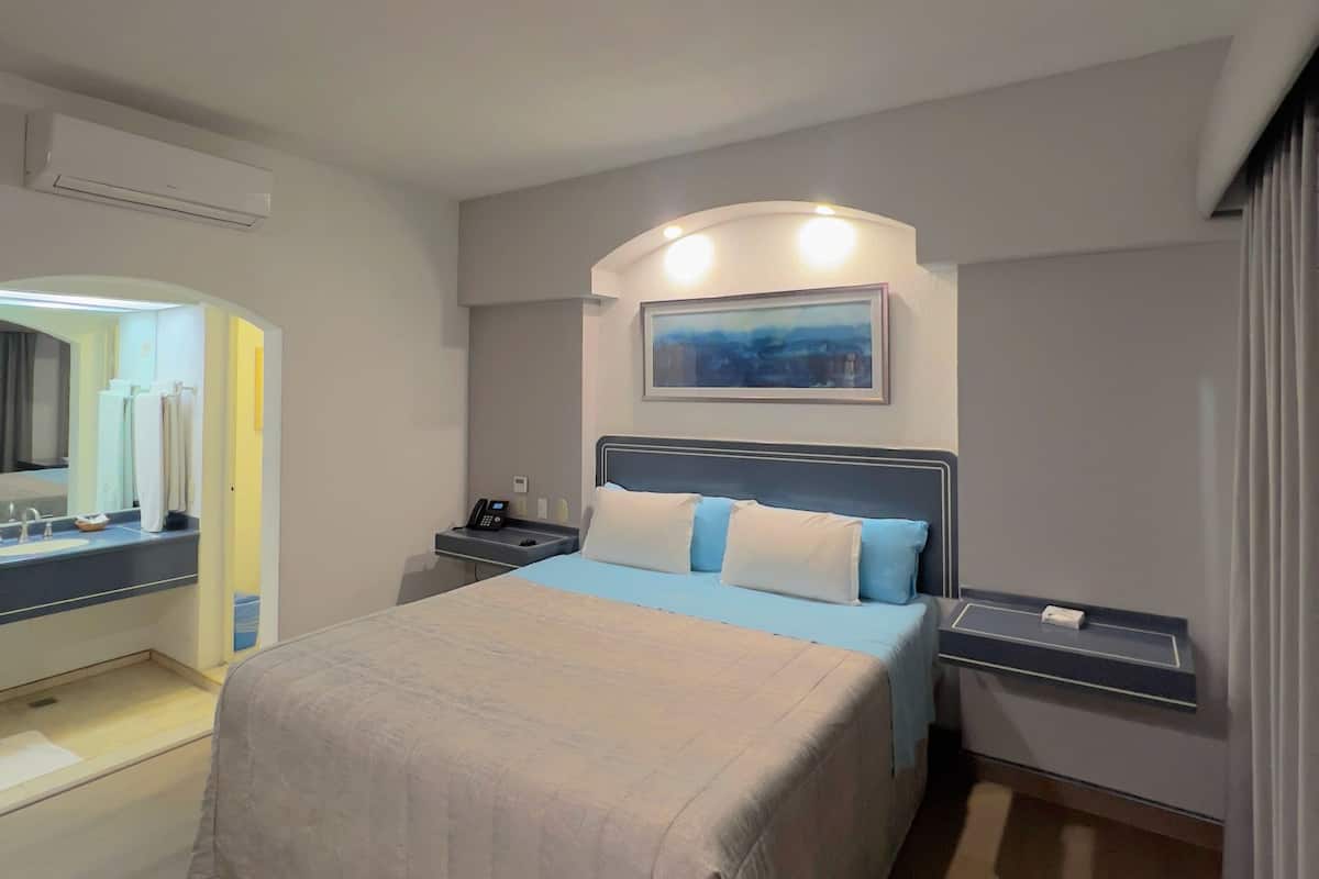 A hotel room with a bed, bedside tables, a mirror, an air conditioning unit, and a painting on the wall.