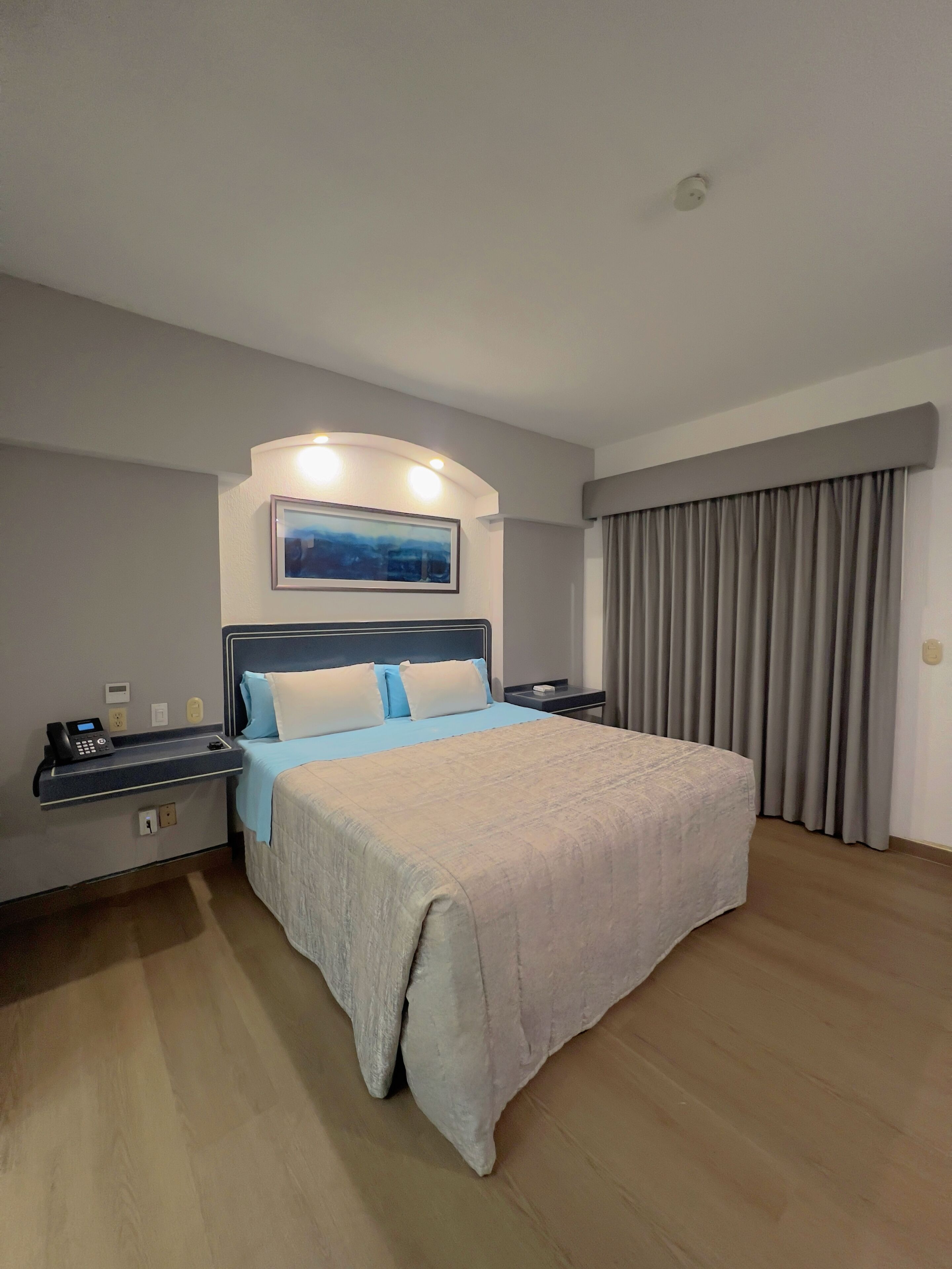 A hotel room with a large bed, a bedside table with a phone, a nightstand, a wall-mounted TV, and a window with curtains.