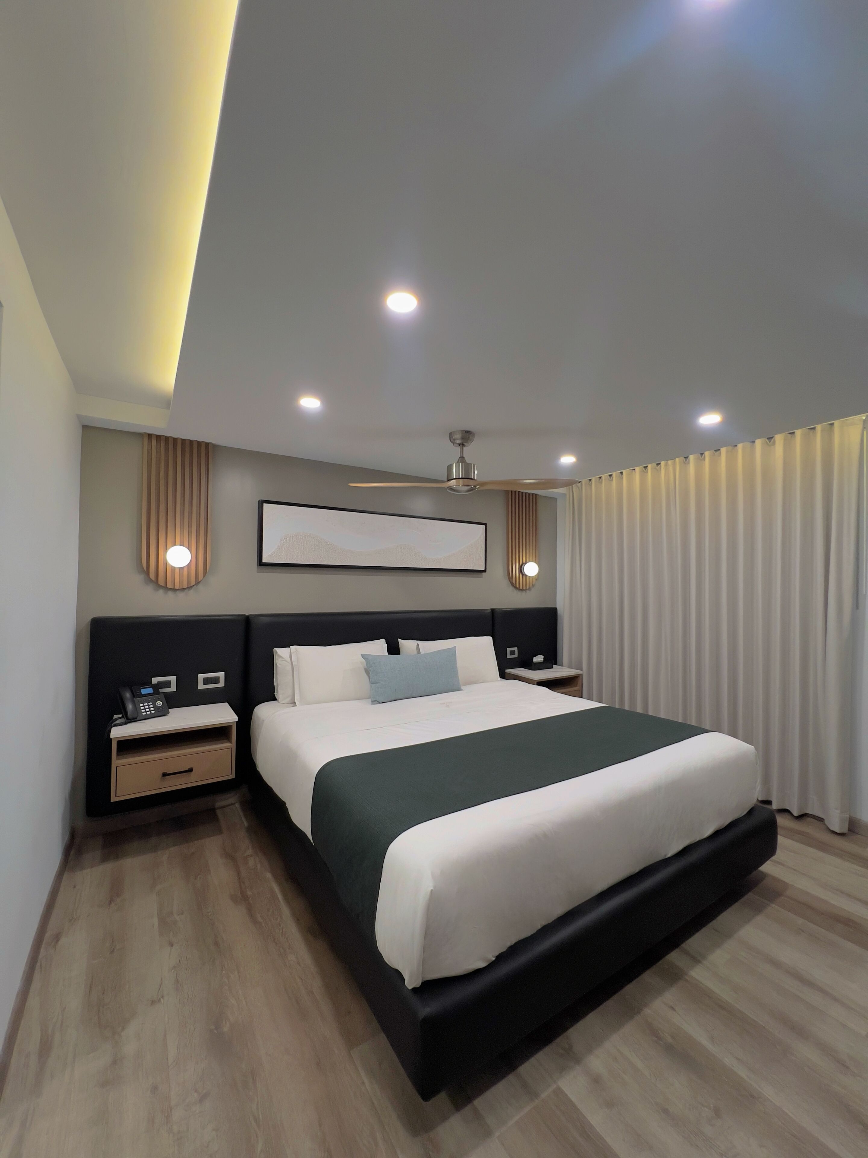 Quarto de hotel moderno com uma cama grande, uma mesa de cabeceira com telefone, ventoinha no teto e iluminação embutida.
