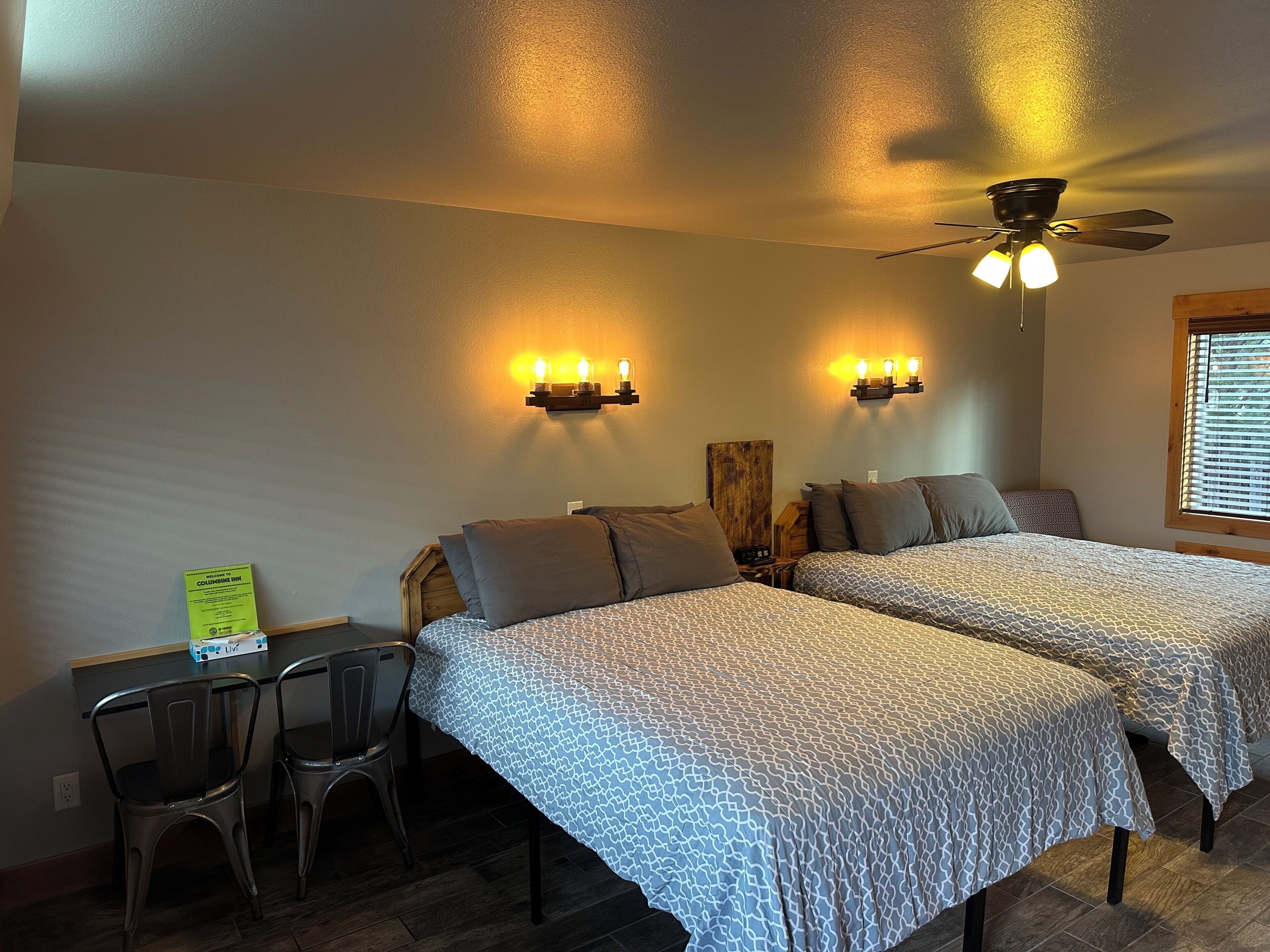 A room with two beds, a desk, chairs, a ceiling fan, and wall-mounted lights.