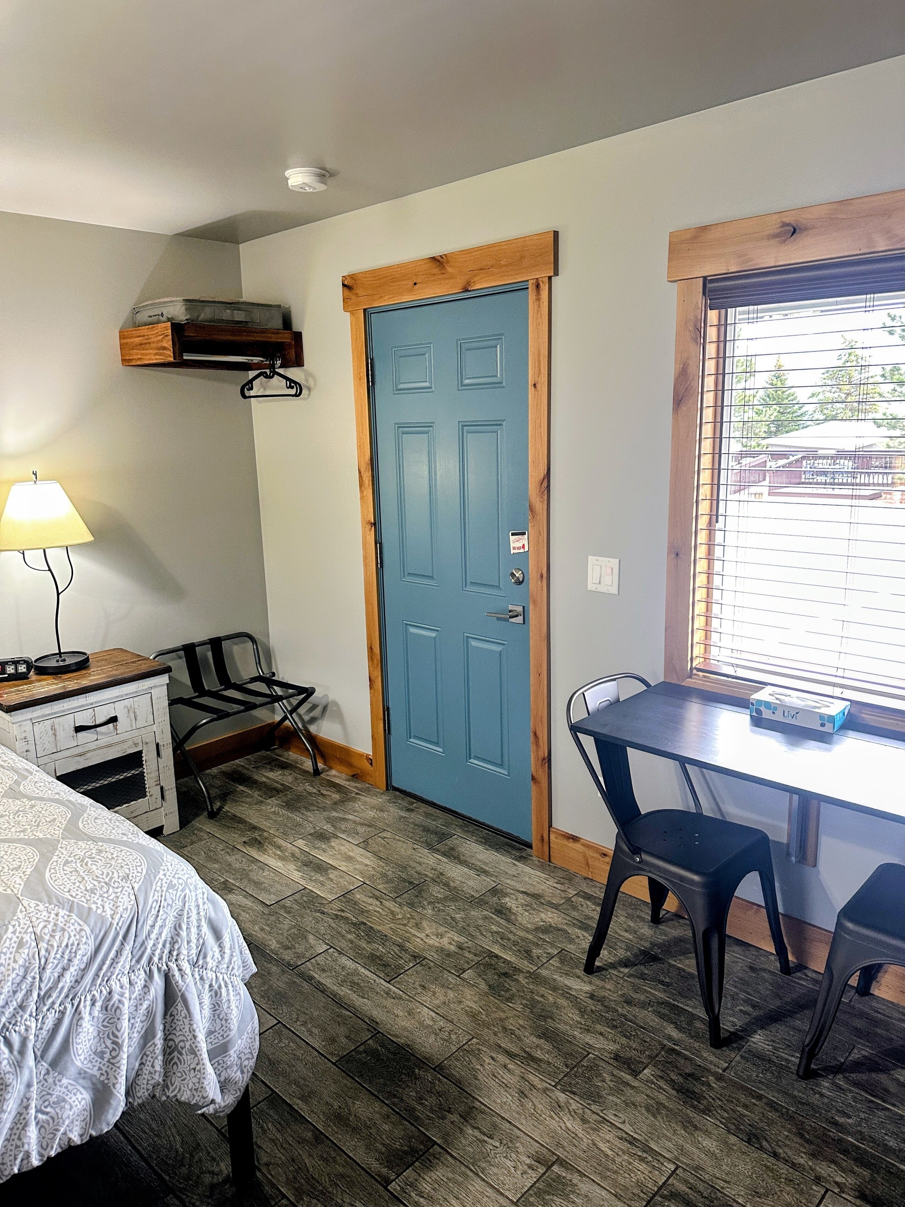 A room with a blue door, a desk with a chair, a bed with a patterned bedspread, a lamp, and a window with blinds.