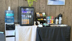 A room with a water dispenser, a refrigerator with beverages, a coffee maker, a microwave, a table with a black tablecloth, and a wall-mounted shelf with snacks. - Town House Motor Inn (Worland)