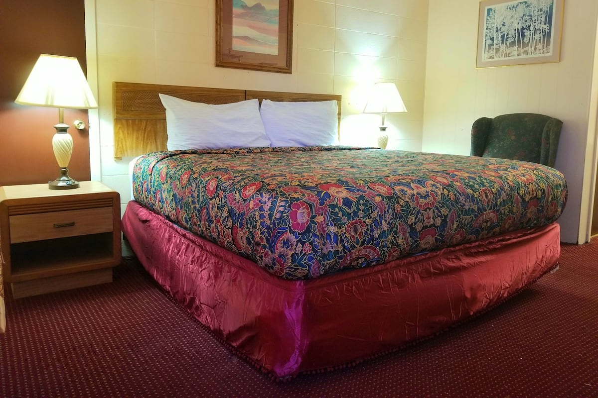 Une chambre d’hôtel avec un grand lit, deux lampes de chevet, une table de nuit en bois, un fauteuil vert et deux tableaux encadrés au mur.