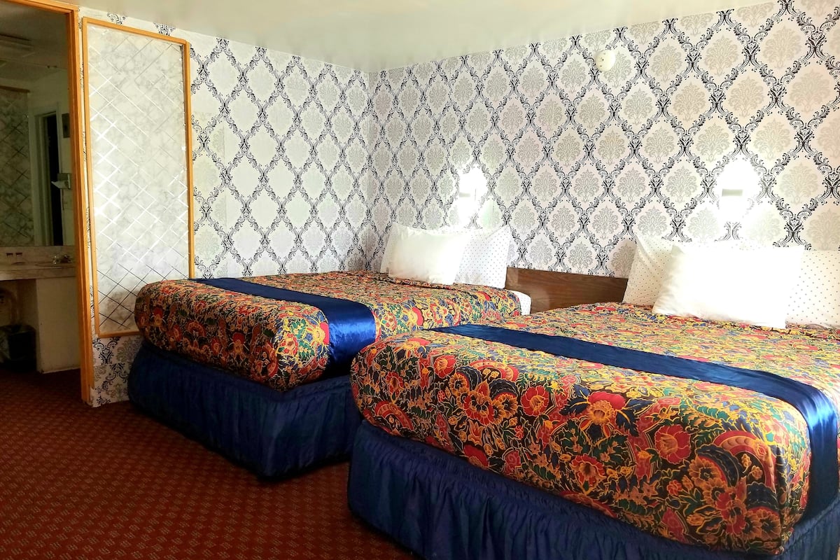 Une chambre d’hôtel avec deux lits, chacun agrémenté de draps à motifs floraux et d’une tête de lit bleue. La chambre est dotée d’un papier peint à motifs et d’une moquette rouge.