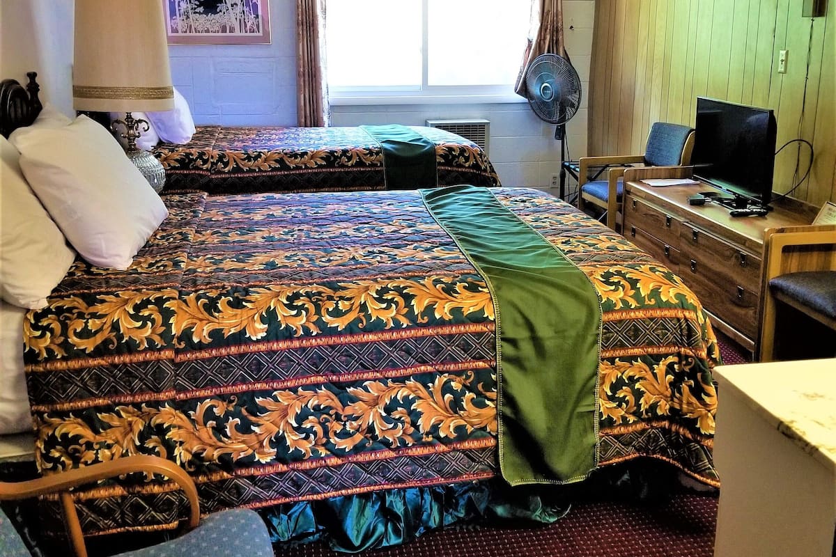 Une chambre d’hôtel avec deux lits, un bureau avec un ordinateur, une chaise, une fenêtre avec des rideaux et un ventilateur fixé au mur.