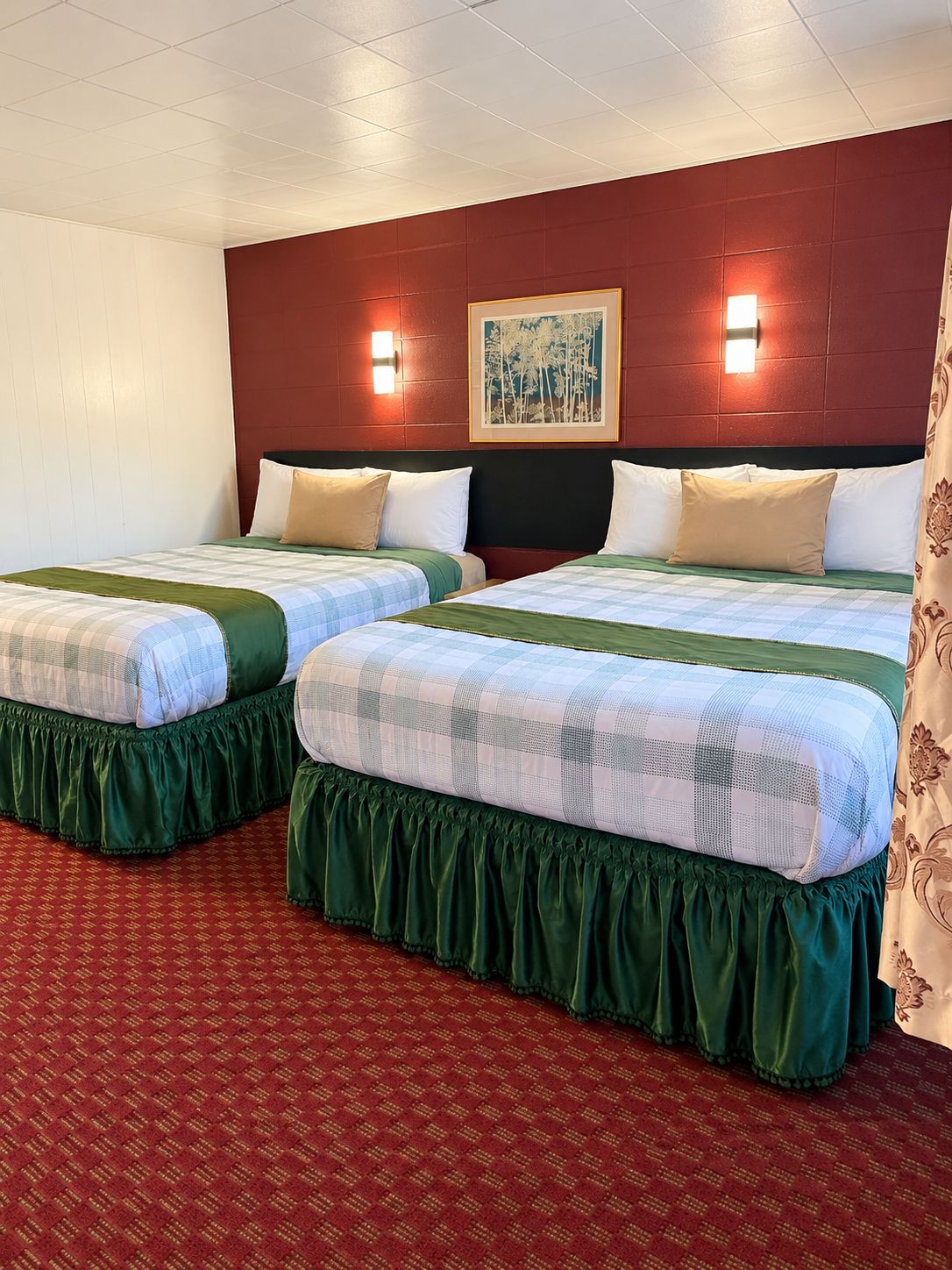 A hotel room with two beds, each with a green skirted bedspread and white pillows. There is a framed picture on the wall between the beds.