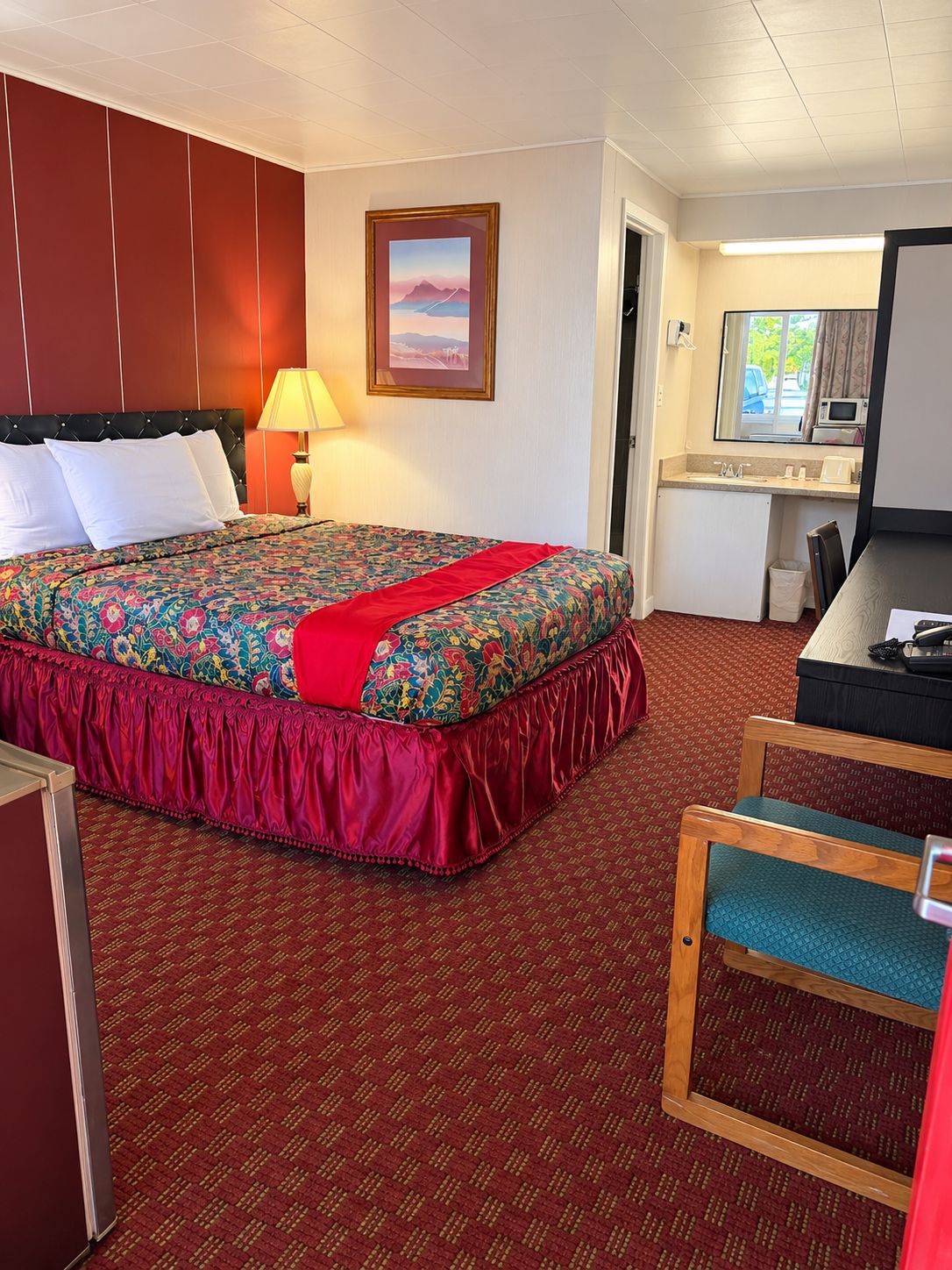 Quarto de hotel com cama, escrivaninha, cadeira, luminária e quadro na parede.