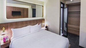 Una habitación de hotel con una cama grande, una mesita de noche con lámpara, un televisor montado en la pared y un armario.