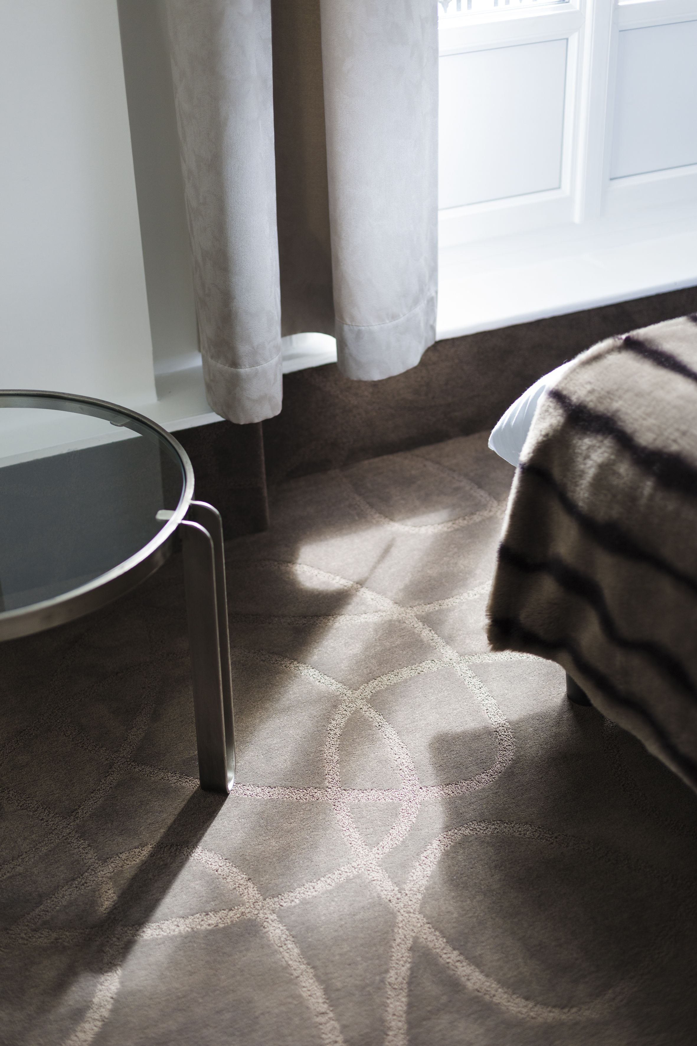 A room with a glass table, a patterned carpet, and a bed with a striped blanket.