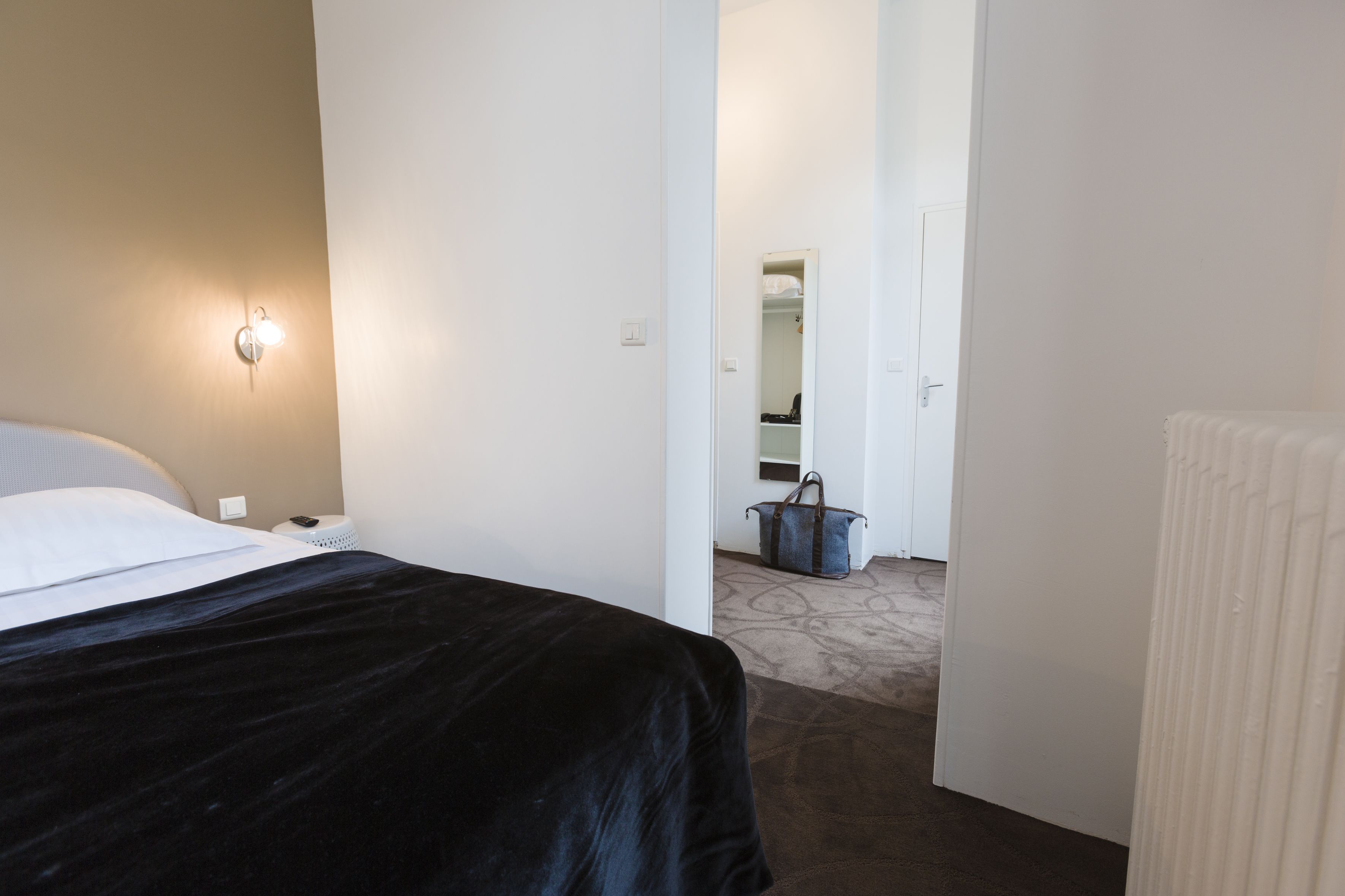 A hotel room with a bed, a nightstand, a lamp, a radiator, and a hallway leading to another room.