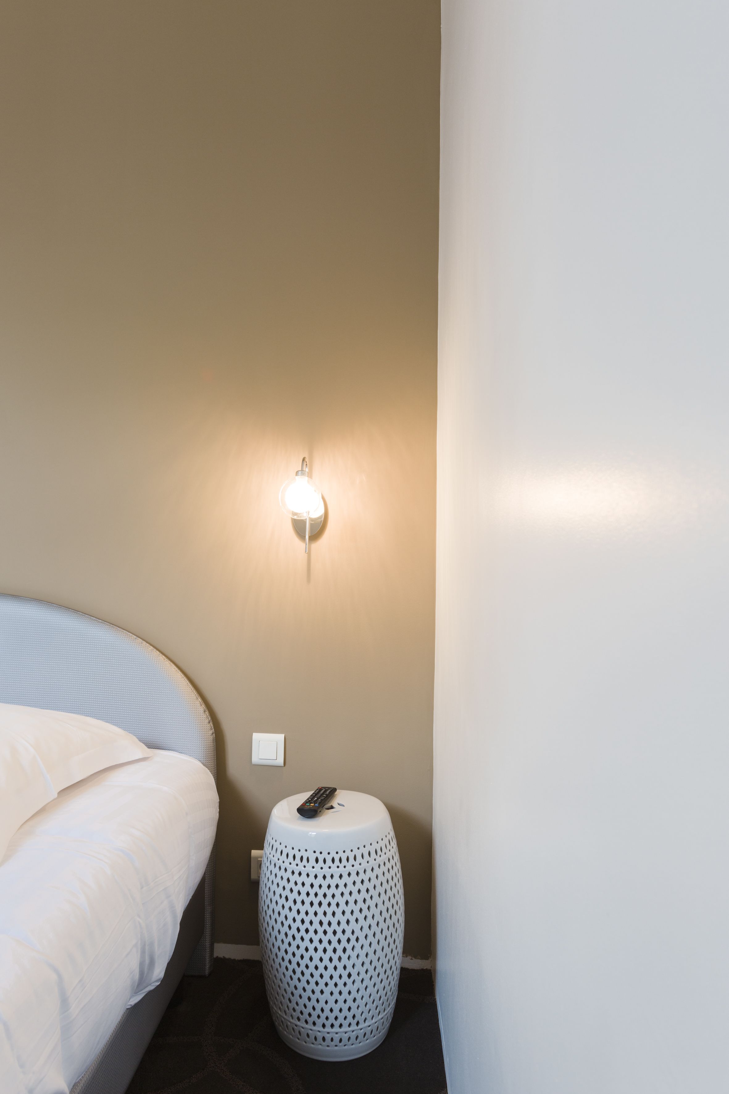 Una cama bien hecha con sábanas blancas, un taburete blanco y redondo con diseño perforado y una lámpara de pared.
