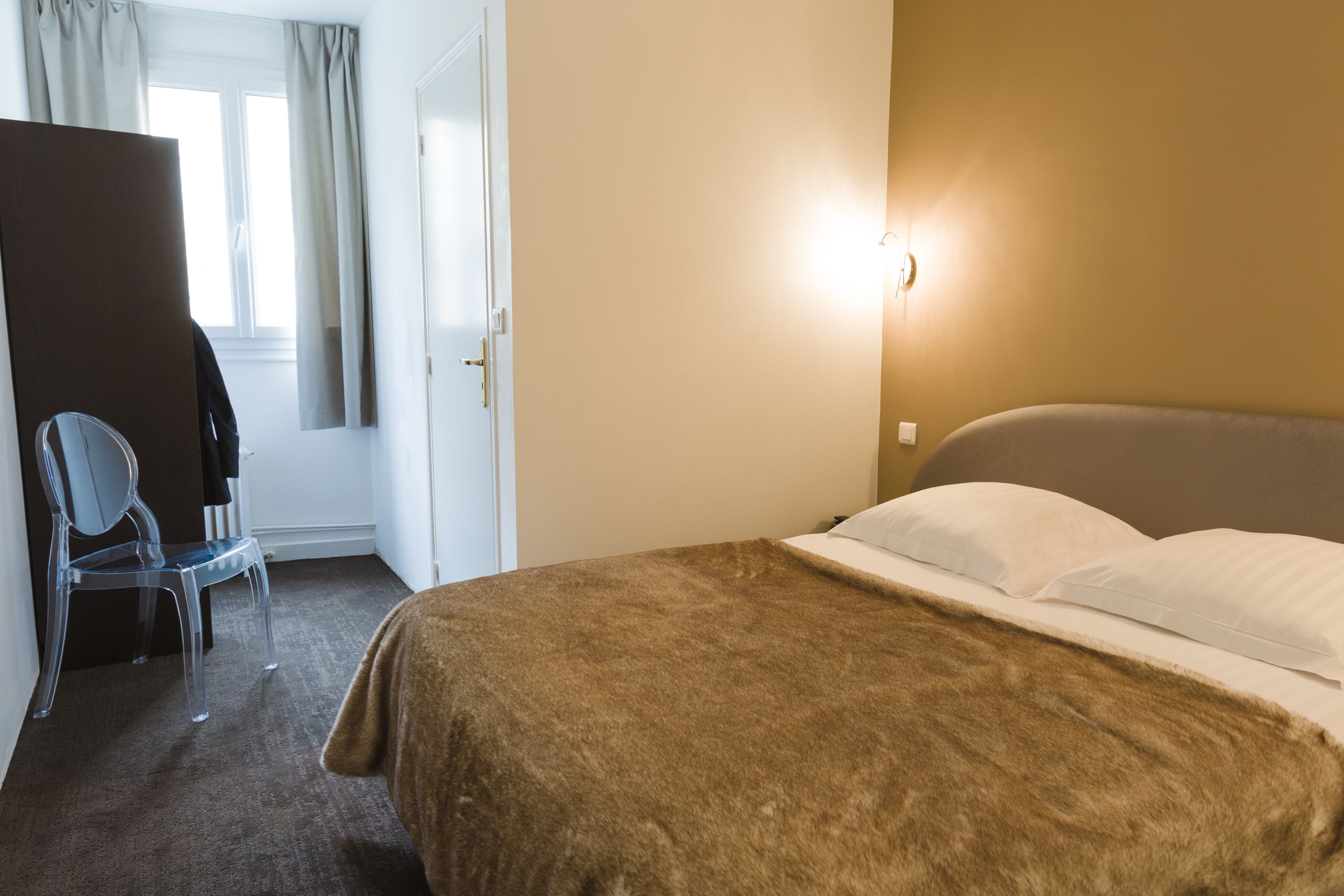 Een hotelkamer met een bed, een stoel, een raam met gordijnen en een wandlamp.
