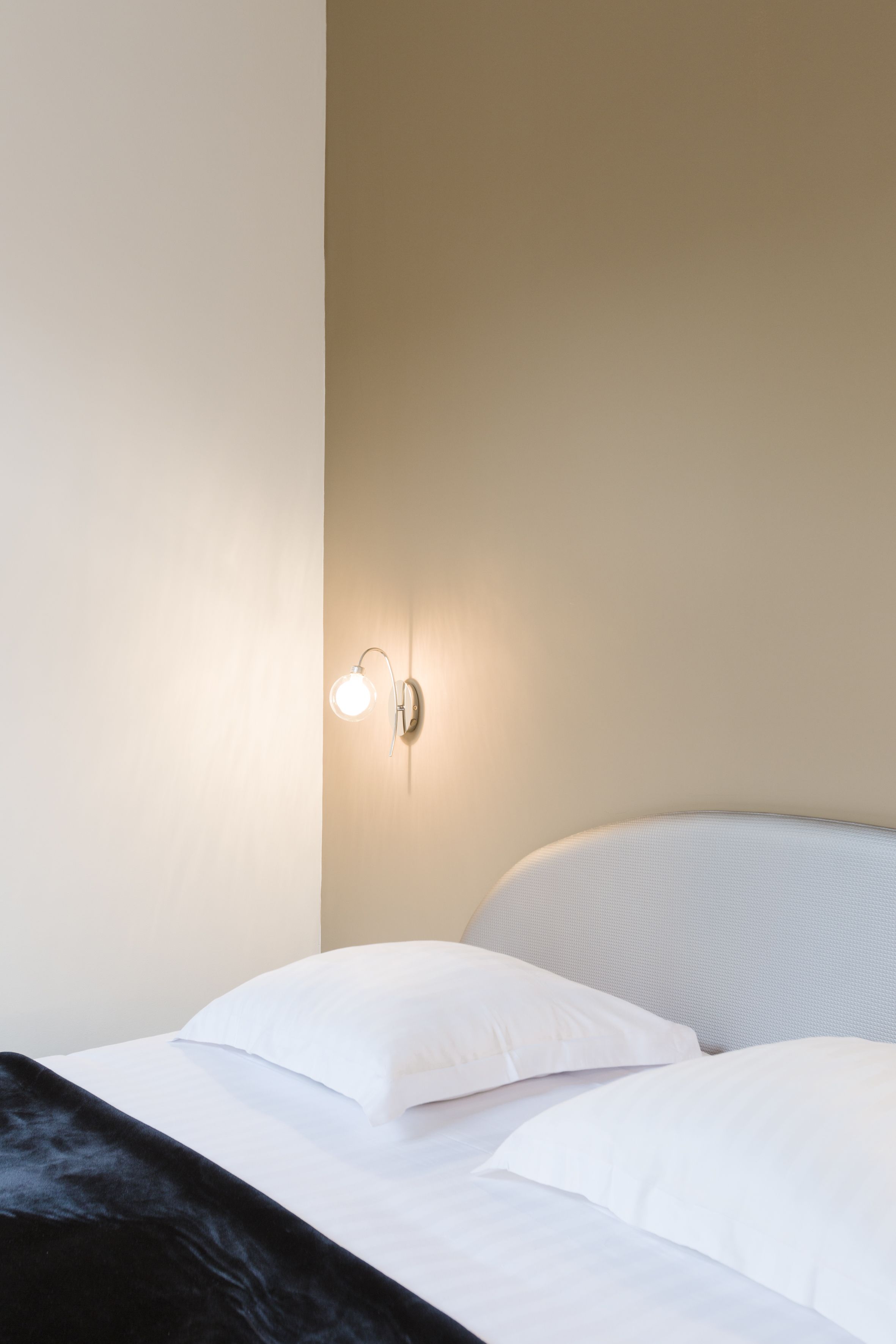 Una cama con ropa de cama blanca y un cabecero negro, un aplique de pared y una pared lisa.