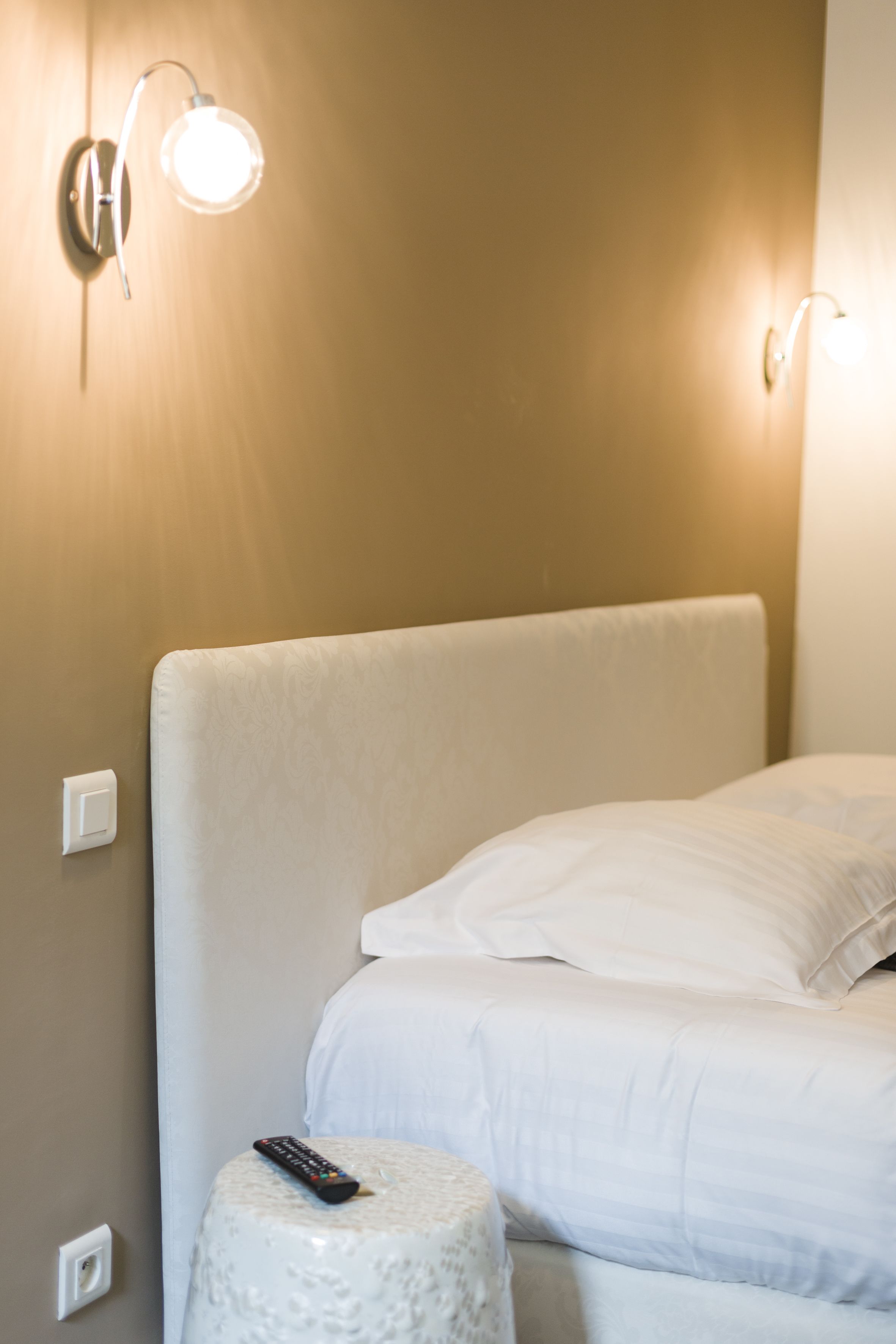 A neatly made bed with white bedding, a remote control on a round stool, and wall-mounted light fixtures.
