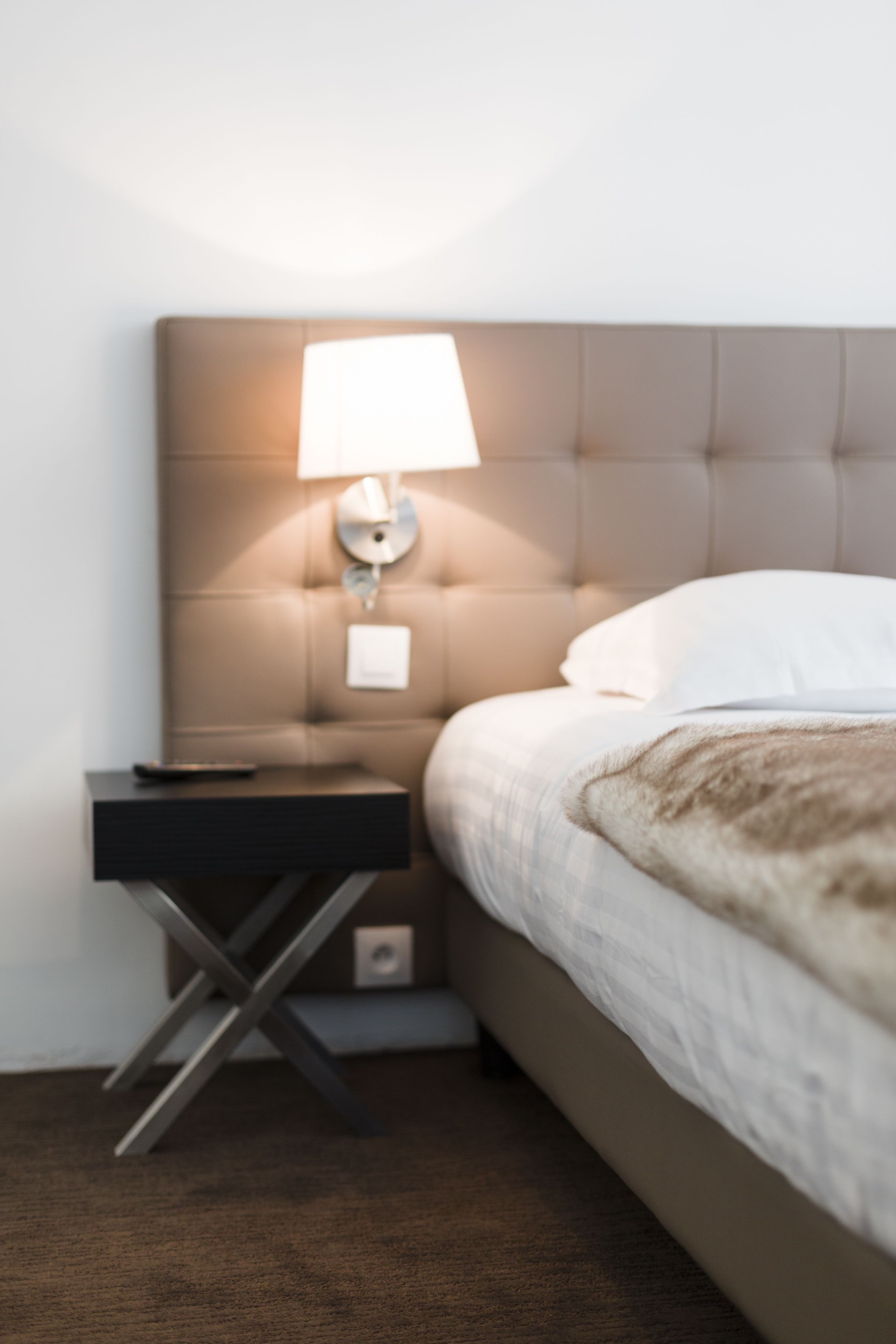 A neatly made bed with a white pillow and a brown throw blanket, a bedside table with a lamp, and a modern, tufted headboard.