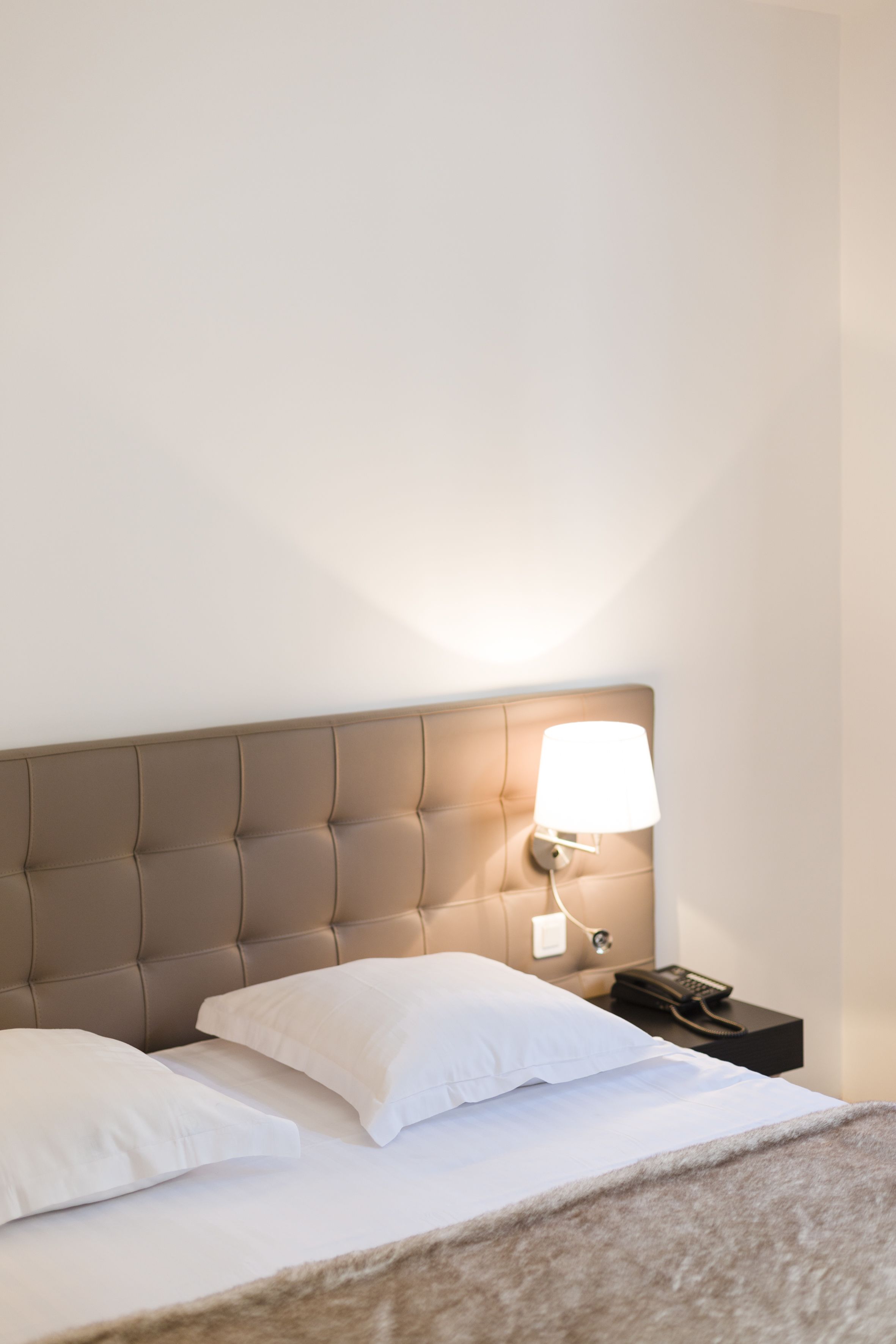 A neatly made bed with white linens and a brown headboard. A bedside lamp is on, and a phone is on the nightstand.